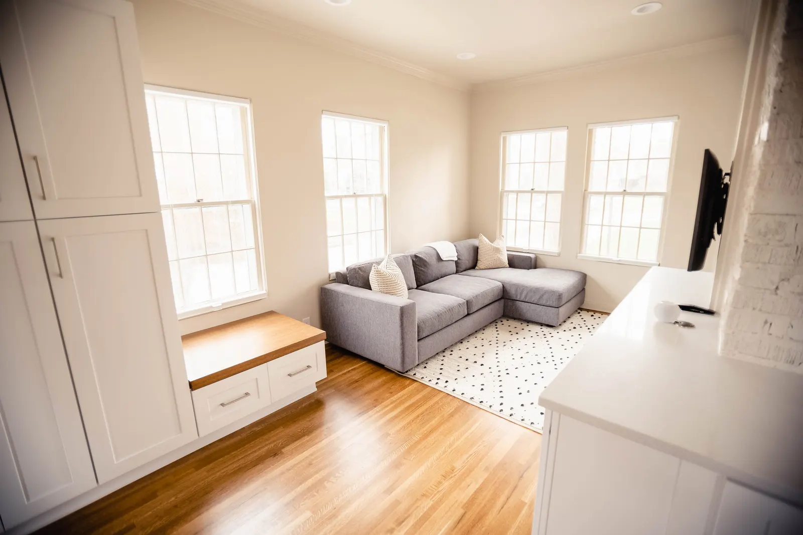 Living room with a gray sectional sofa, white walls, and wooden floor. Four windows above the sofa, a patterned rug, and a white cabinet with a wooden top to the left.