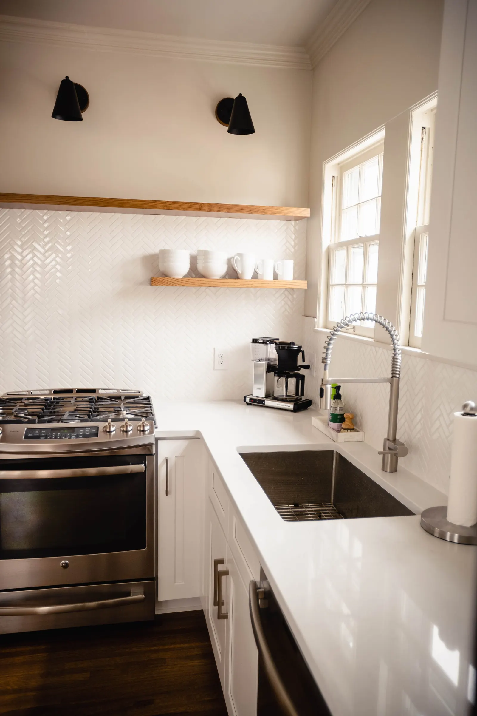 A modern kitchen with white cabinets, stainless steel stove, and a black sink under a window. Two black wall sconces hang above, and floating wooden shelves hold white bowls and mugs. Kitchen appliances like a coffee maker and grinder are on the coun