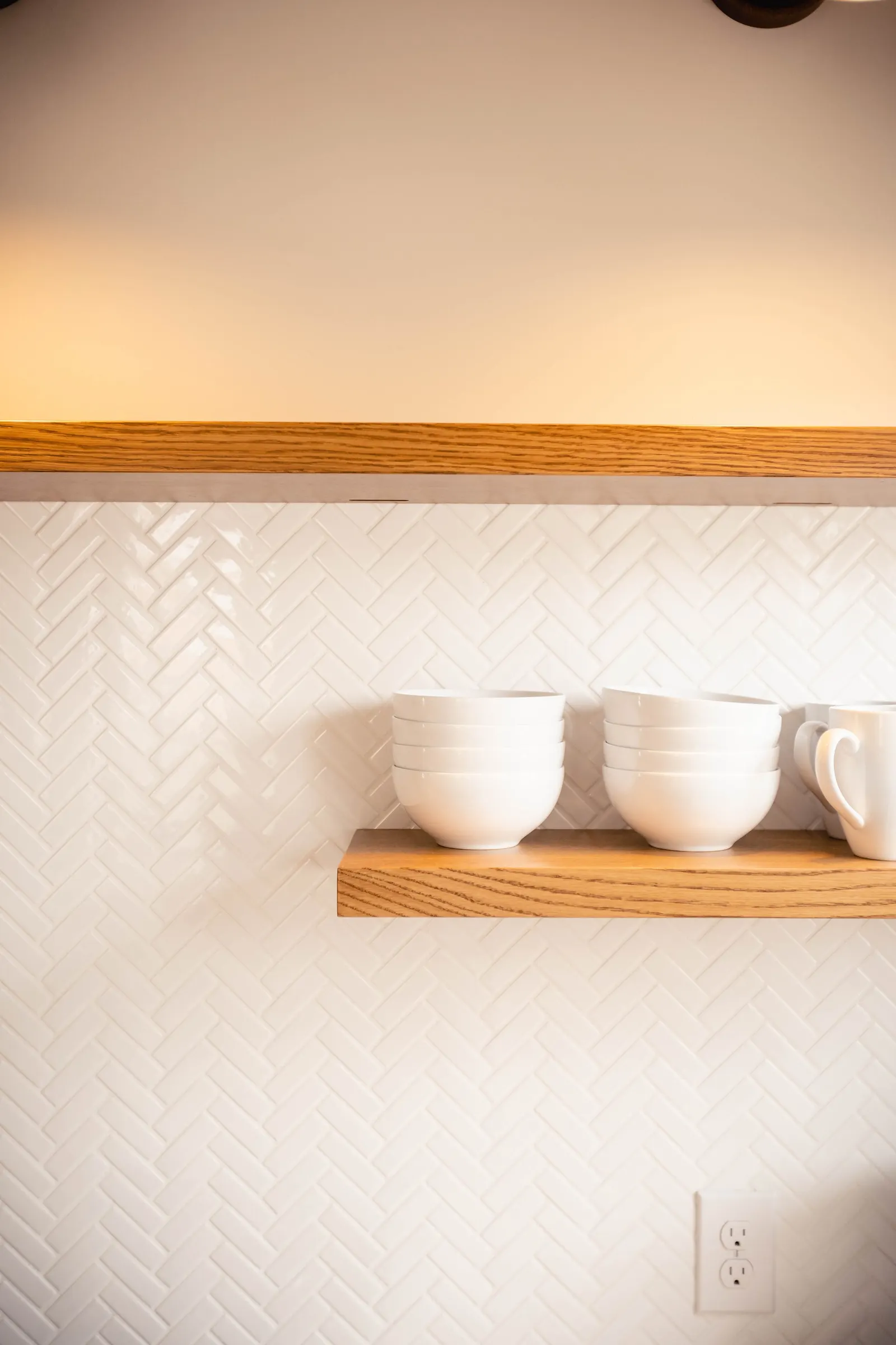 A kitchen wall with a herringbone white tile backsplash, two wooden floating shelves, and white bowls and a mug on the lower shelf.