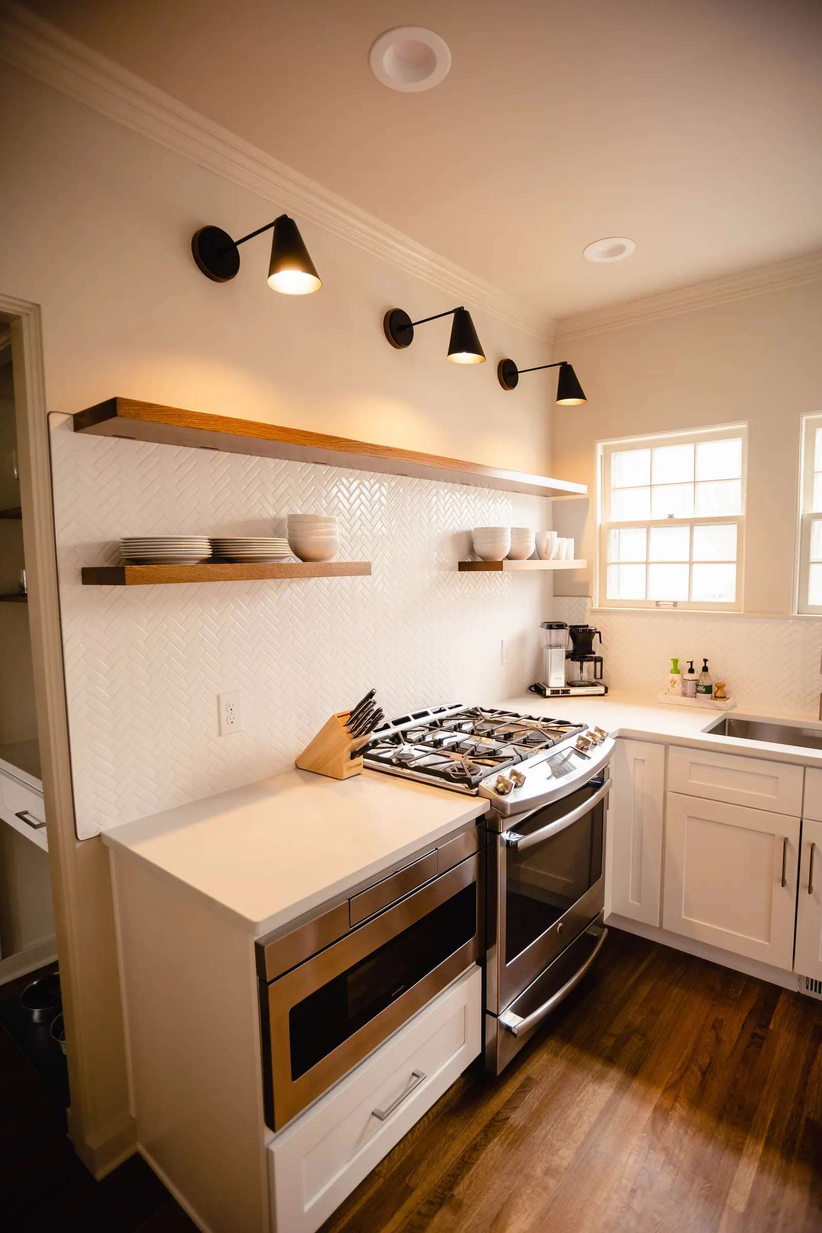Modern kitchen with white cabinets, a stainless steel oven, and white wall tiles with a herringbone pattern. Open wooden shelves hold bowls and plates, black wall-mounted lights, and natural light from two windows.