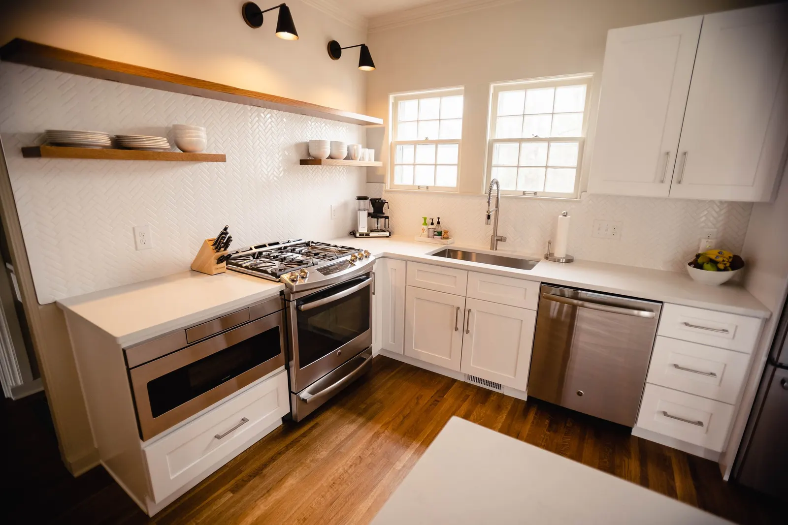 Modern kitchen with white cabinets, stainless steel appliances, and wooden accents. Open shelving with dishes, a gas stove, a coffee maker, and a bowl of fruit on the counter. Two windows above the sink, hardwood flooring.