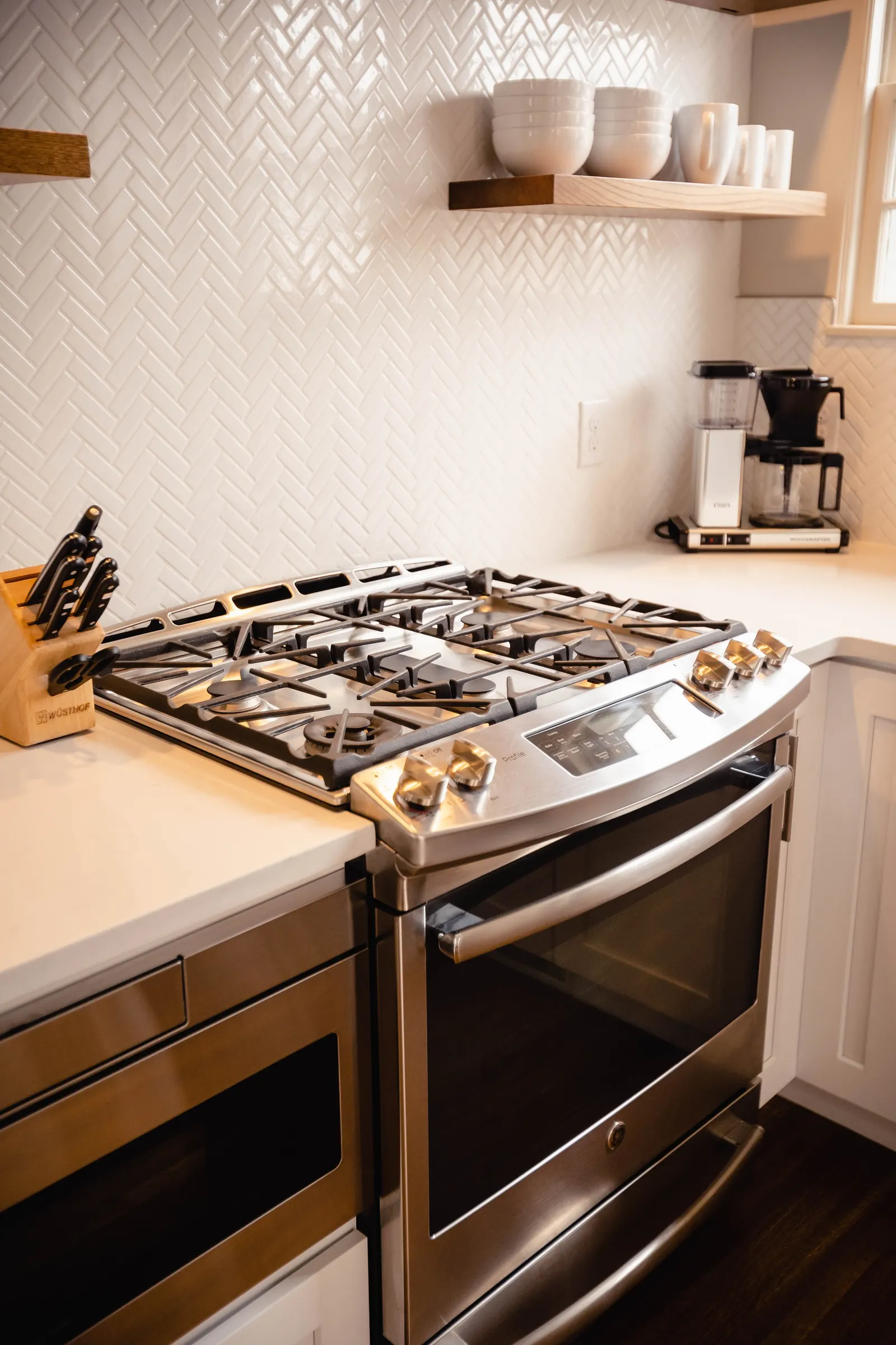 A modern kitchen with a stainless steel gas stove, a knife block on the left, a wooden shelf with white bowls and cups above, and a coffee maker with a grinder on the right.