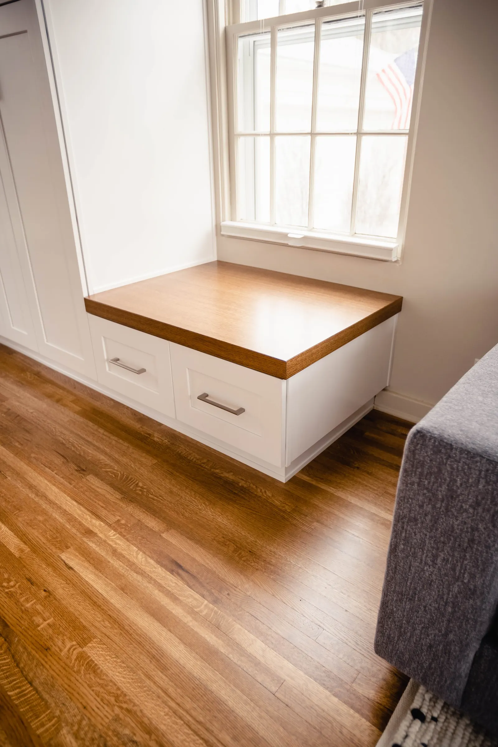Wooden window seat with two drawers below, next to a window with an American flag banner outside.