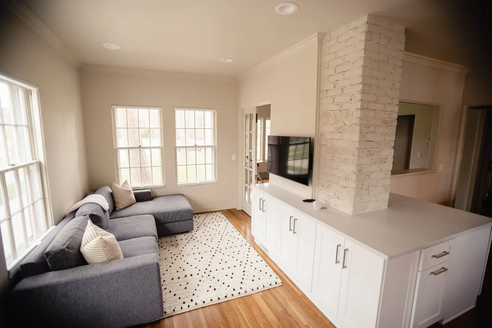Living room with a gray sectional sofa, white cabinets, a wall-mounted TV, white brick column, hardwood floor, three large windows, and a white polka dot rug.