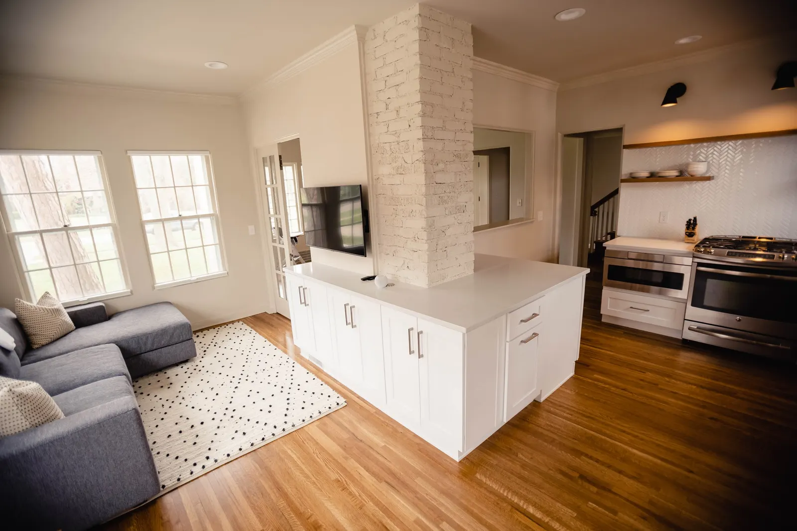 Open-concept living room and kitchen with hardwood floors, white cabinets, brick column, gray sofa, large windows, wall-mounted TV, and modern appliances.