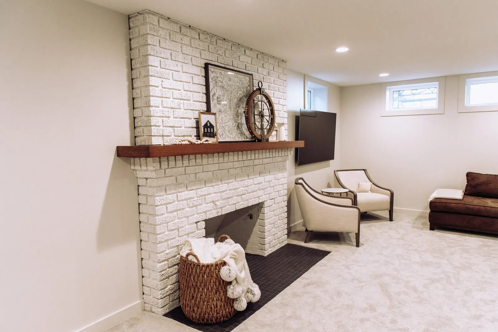 Living room with white brick fireplace, beige carpet, two windows, a television, beige armchair, brown sofa, and decorative items on the mantel.