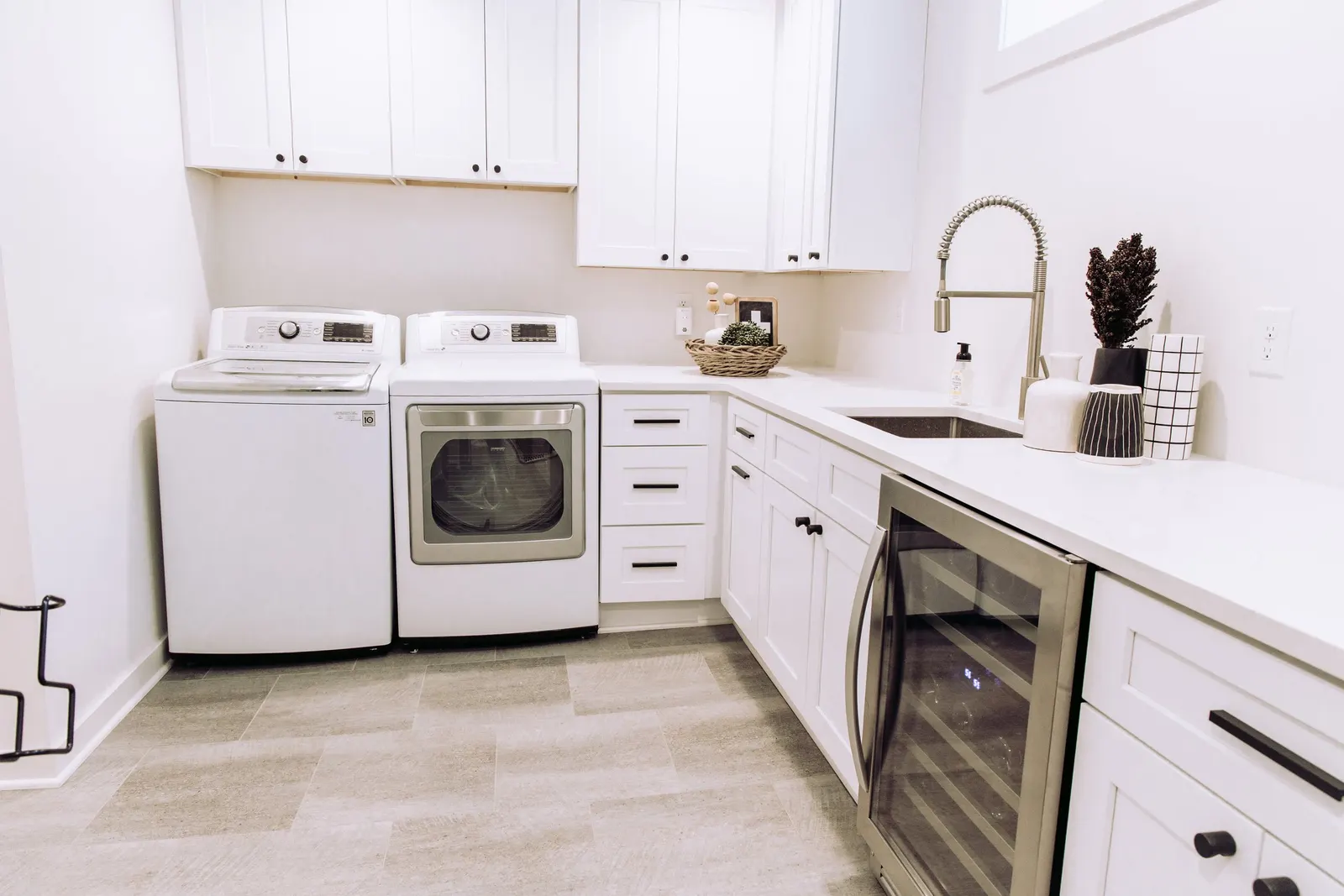 1922 Basement Remodel, laundry room, white cabinets, matte black hardware, brushed nickel faucet, under mount sink, tile floors, beverage fridge, custom cabinetry, Lincoln Nebraska, professional contractors, craftsmanship, storage solutions.
