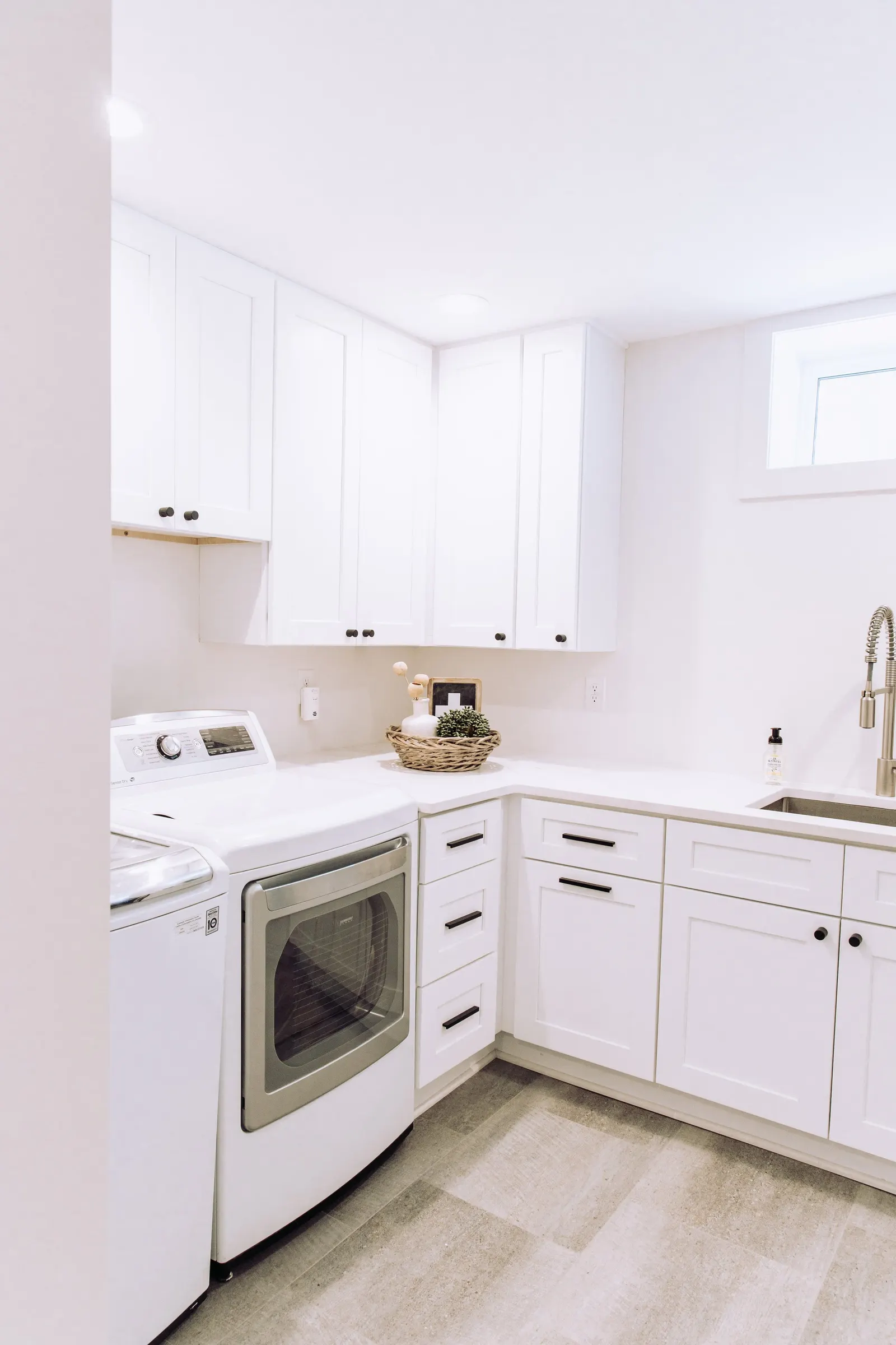 1922 Basement Remodel, laundry room, white cabinets, matte black hardware, brushed nickel faucet, under mount sink, tile floors, beverage fridge, custom cabinetry, Lincoln Nebraska, professional contractors, craftsmanship, storage solutions.