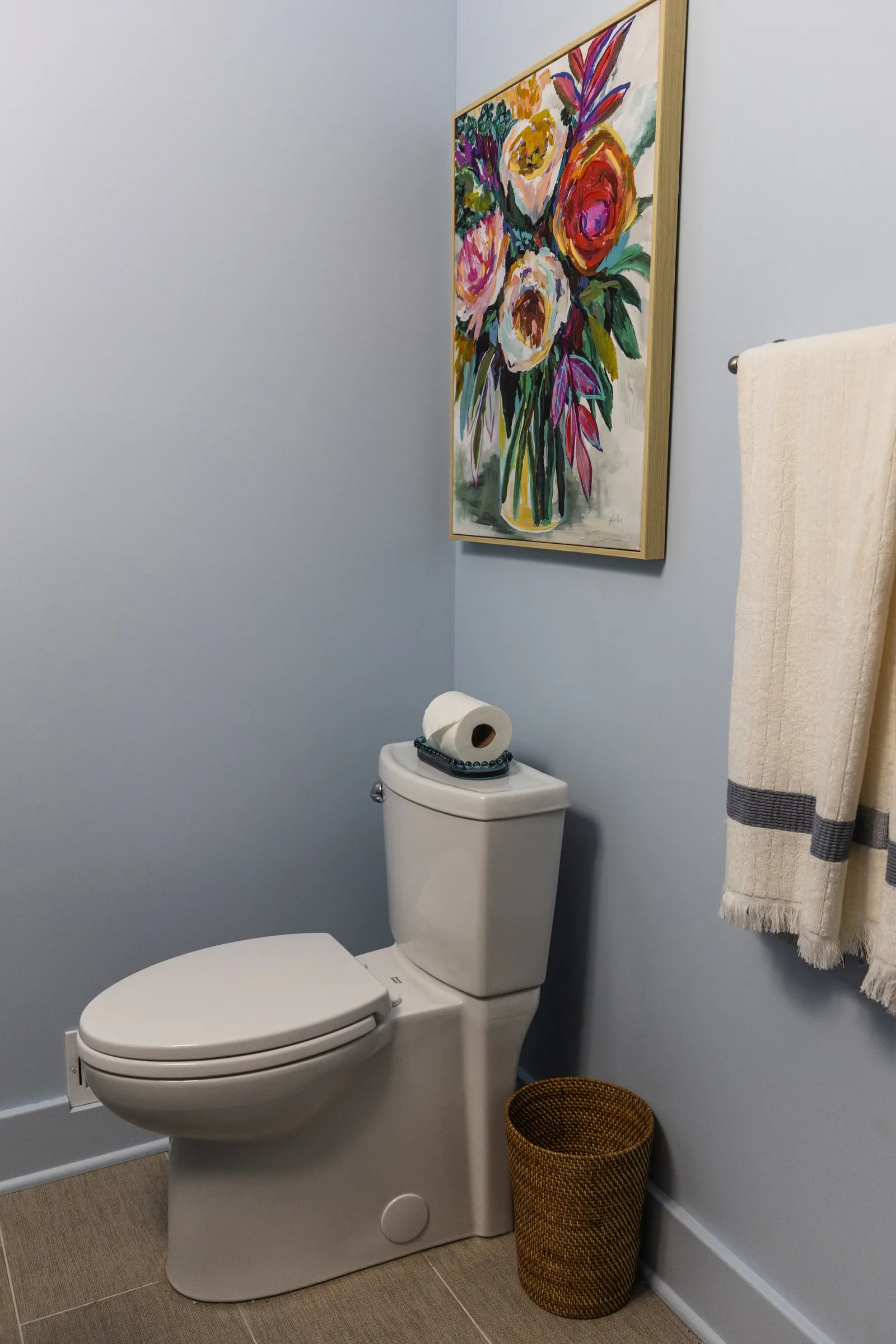 A bathroom with light blue walls, a white toilet with a roll of toilet paper on top, a colorful floral painting hanging on the wall, a beige towel hanging on a towel rack, and a woven wastebasket on the floor.