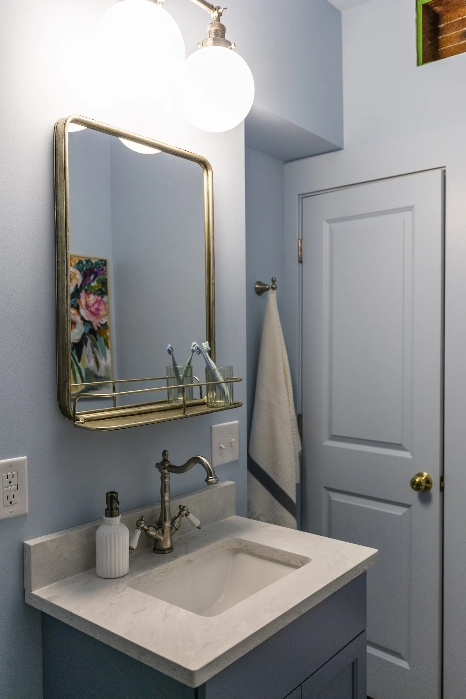 Bathroom vanity with marble countertop, single sink, brass faucet, white ceramic soap dispenser, mirror, wall-mounted light fixture, towel on hook, door with brass knob, and artwork partially visible.
