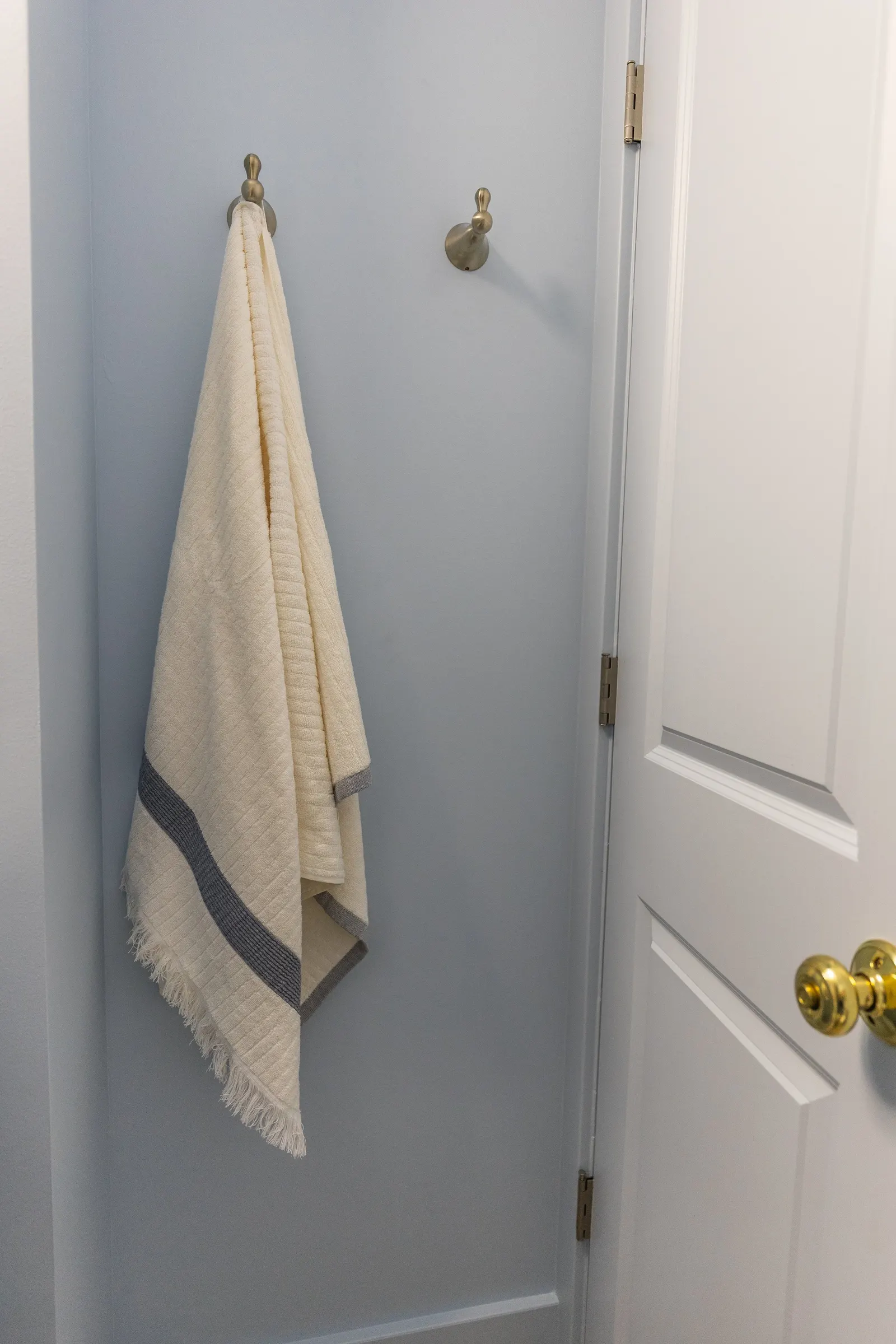Bathroom corner with two brushed metal hooks on a light blue wall, one holding a folded cream towel with a gray border, next to a white door with a gold doorknob.