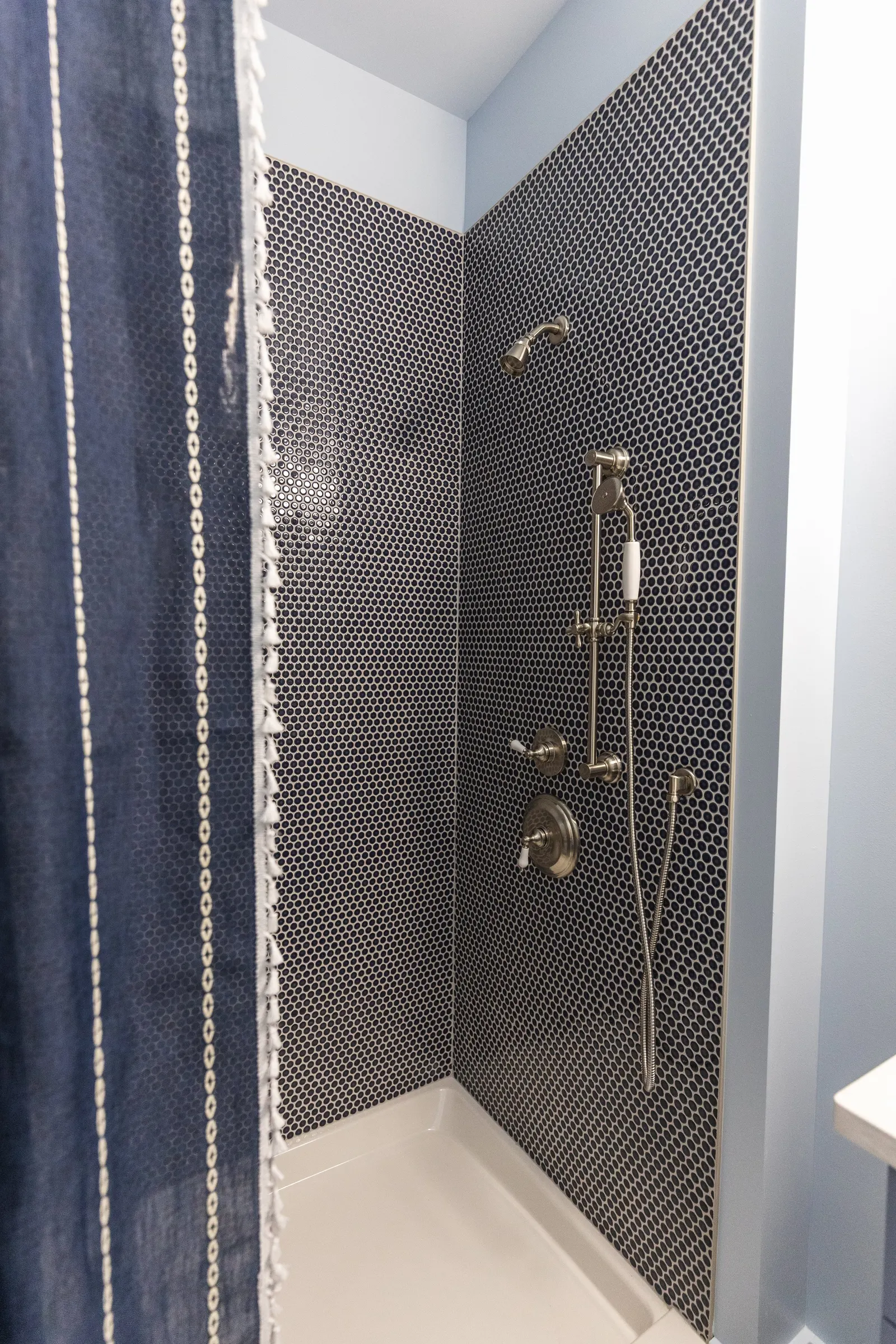A modern shower with black and white mosaic tile walls, featuring a fixed shower head, a handheld shower, and a control panel. A navy blue shower curtain with white lace trim partially covers the shower.