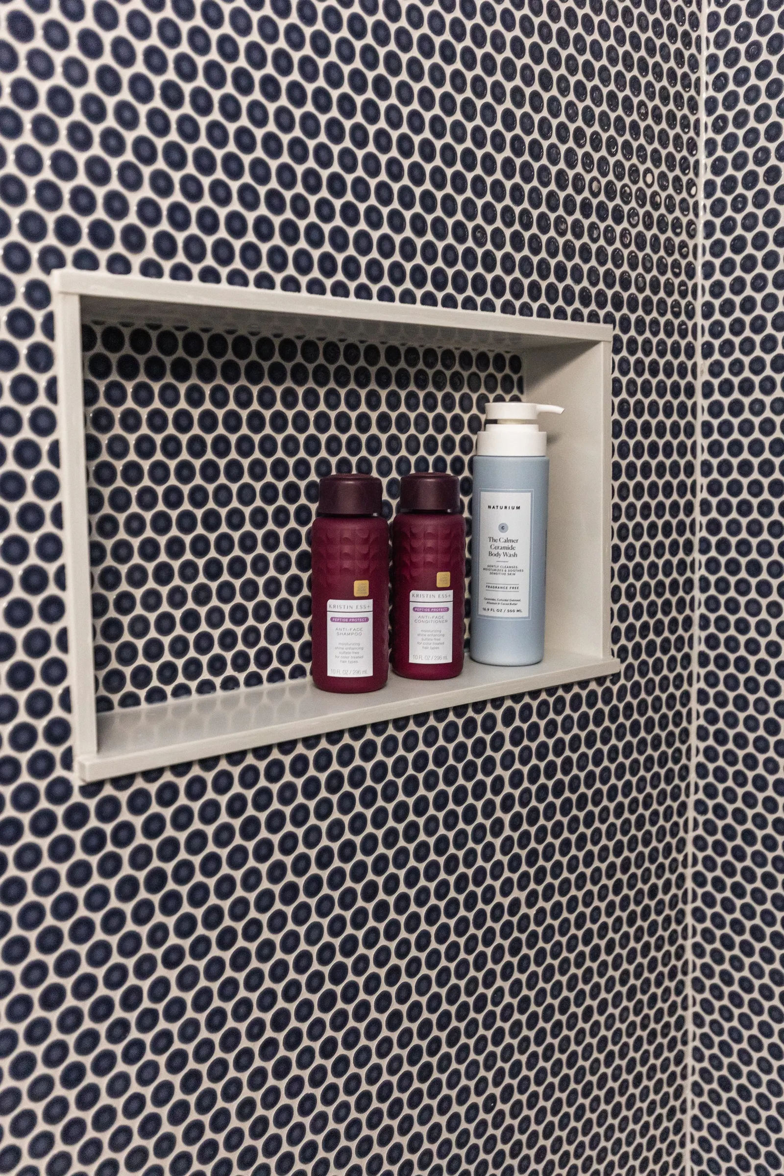 Bathroom shower shelf with two red bottles of Kristin Ess hair products and a blue bottle of Naturium body wash on mosaic black and white tiled wall.