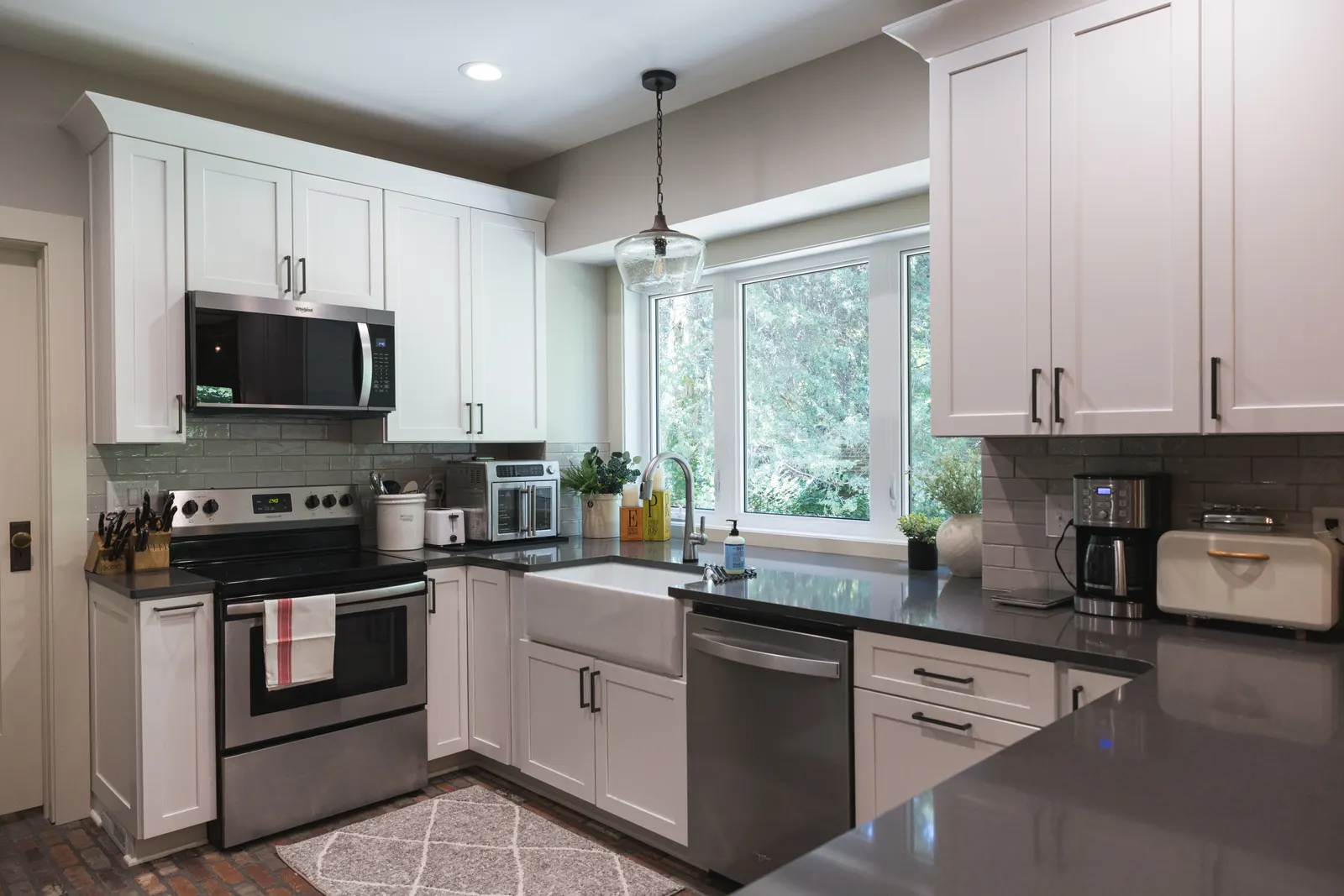 Wide view of the finished kitchen with white shaker cabinets, a stainless range and microwave, a bay window, and a thin brick floor.