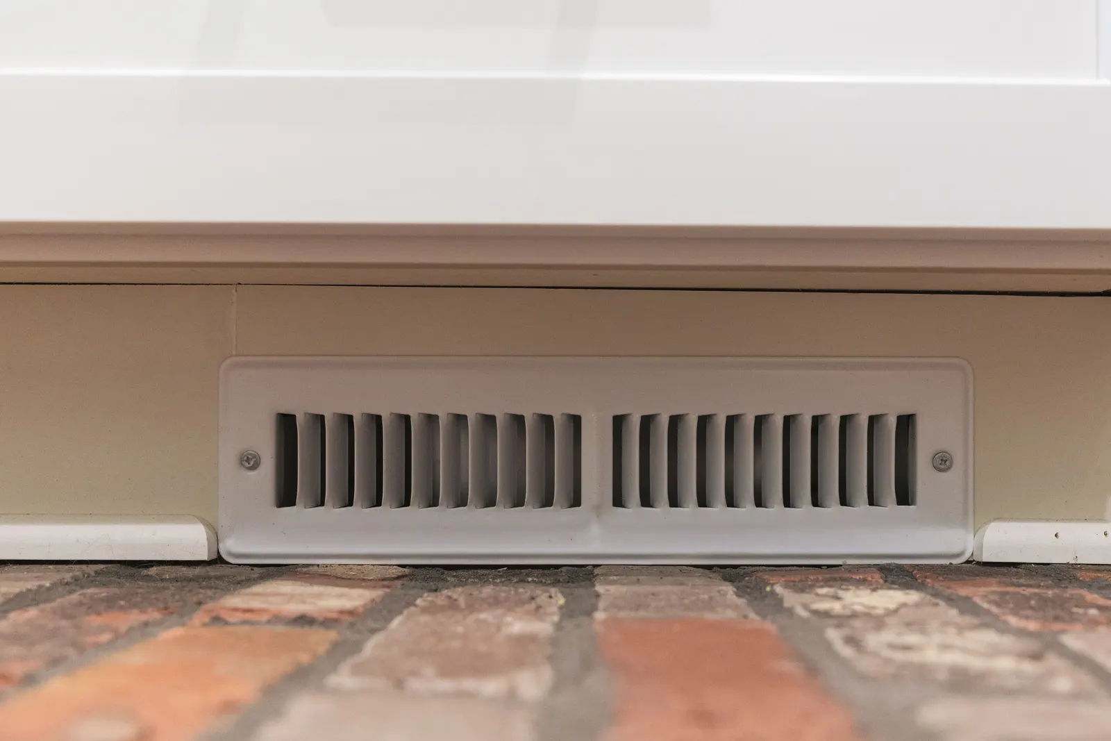 A floor vent cut into the base of a white cabinet above the brick floor.