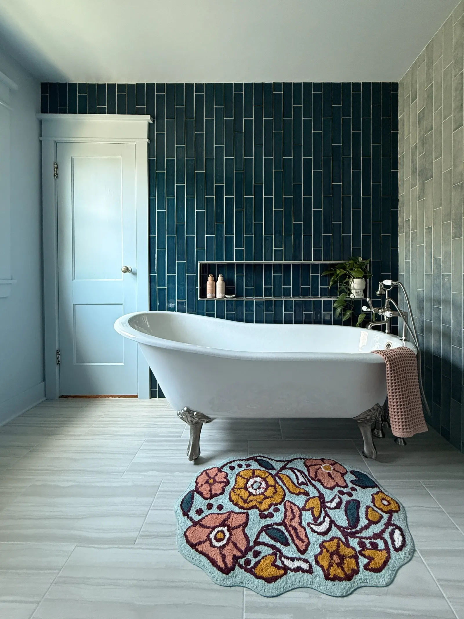 A bathroom with a freestanding bathtub in front of a dark blue tiled wall, a small shelf with bath products, a pink towel on the tub, and a colorful floral rug on the floor.