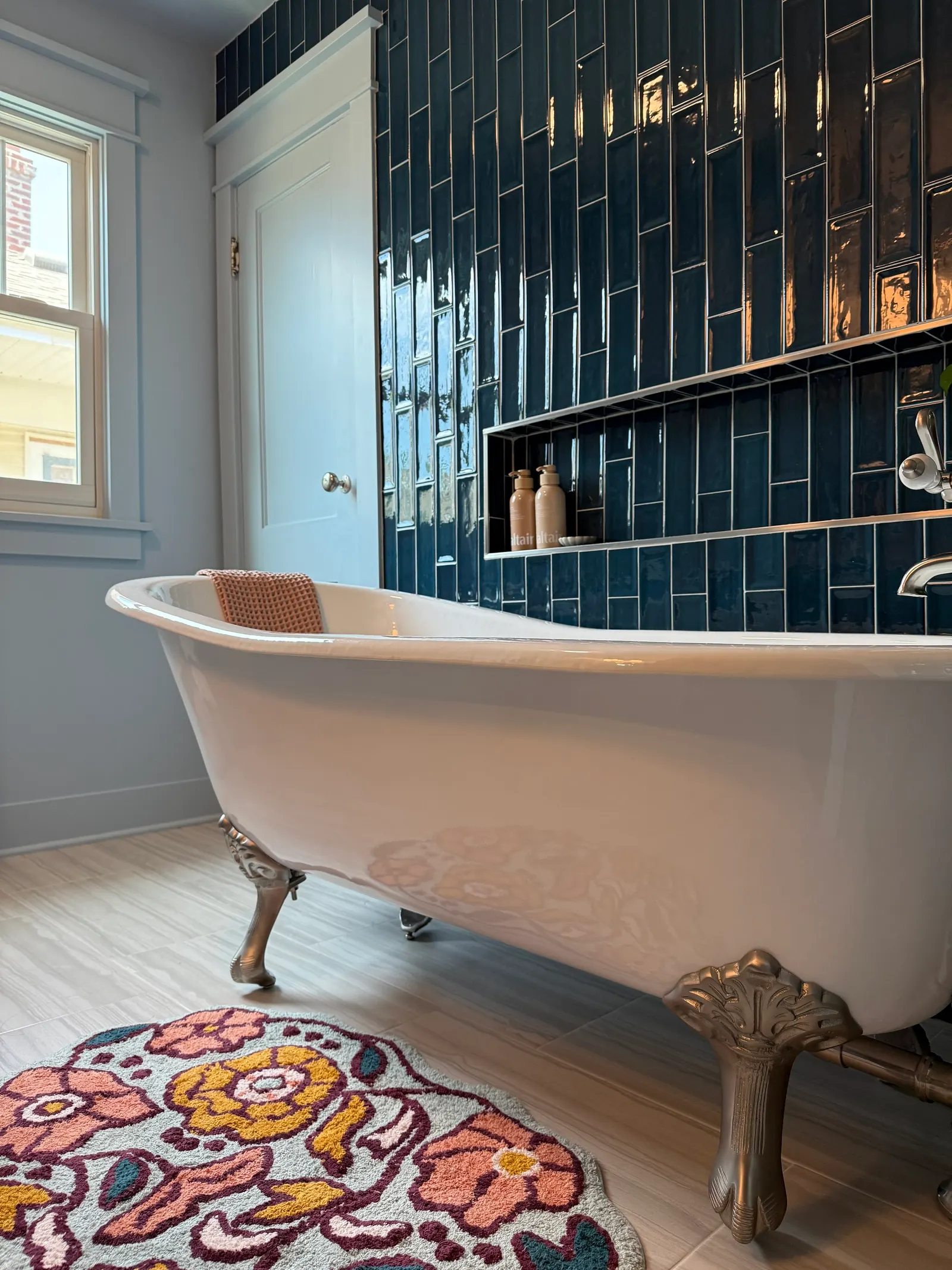 Free-standing clawfoot bathtub with metallic ornate legs, positioned on a floral patterned rug, in a bathroom with blue tiled wall and a white window.