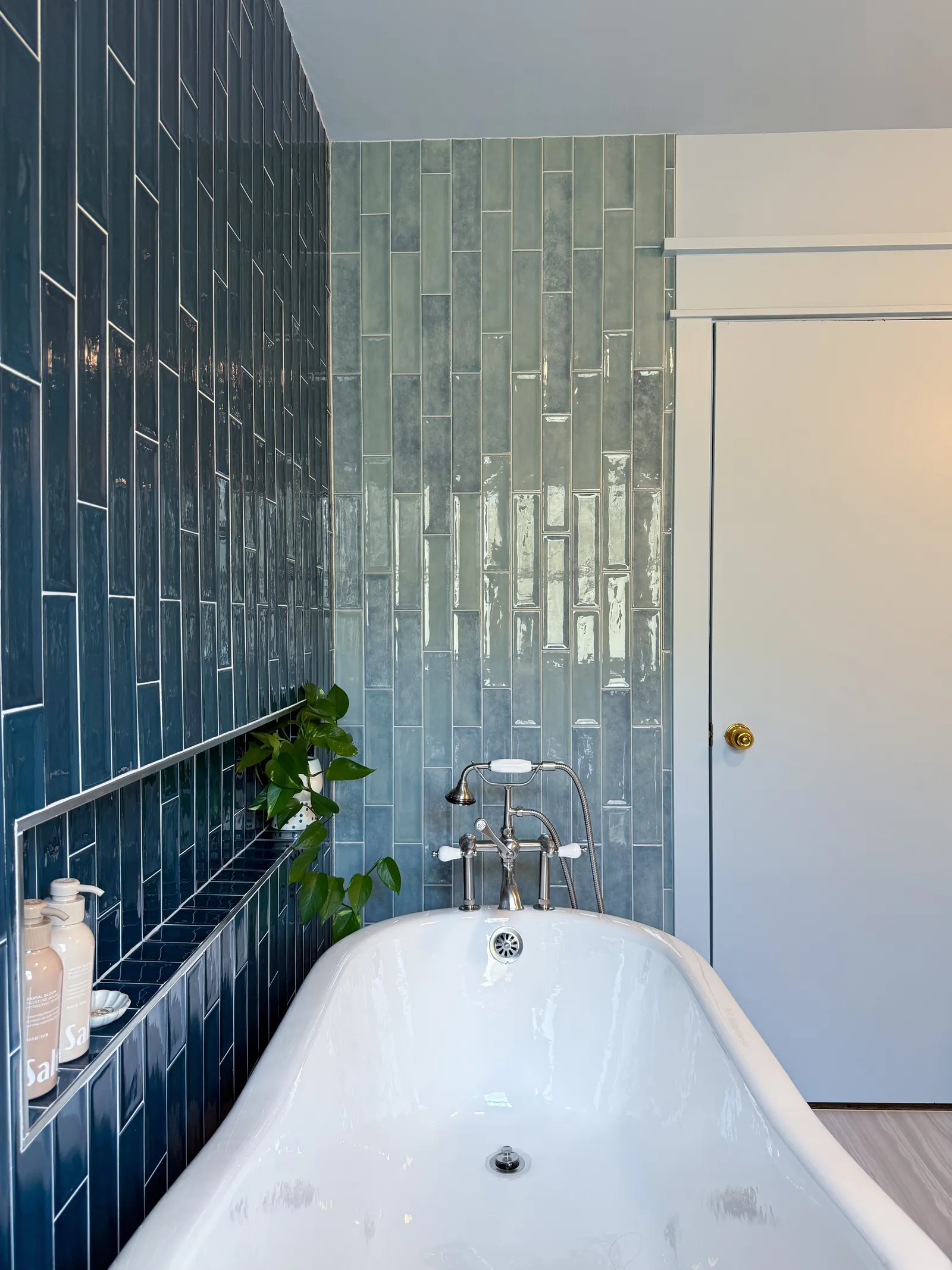 Bathroom with a white clawfoot bathtub, blue tiled wall, green tiled wall, a potted plant, and bottles of soap or lotion on a shelf.