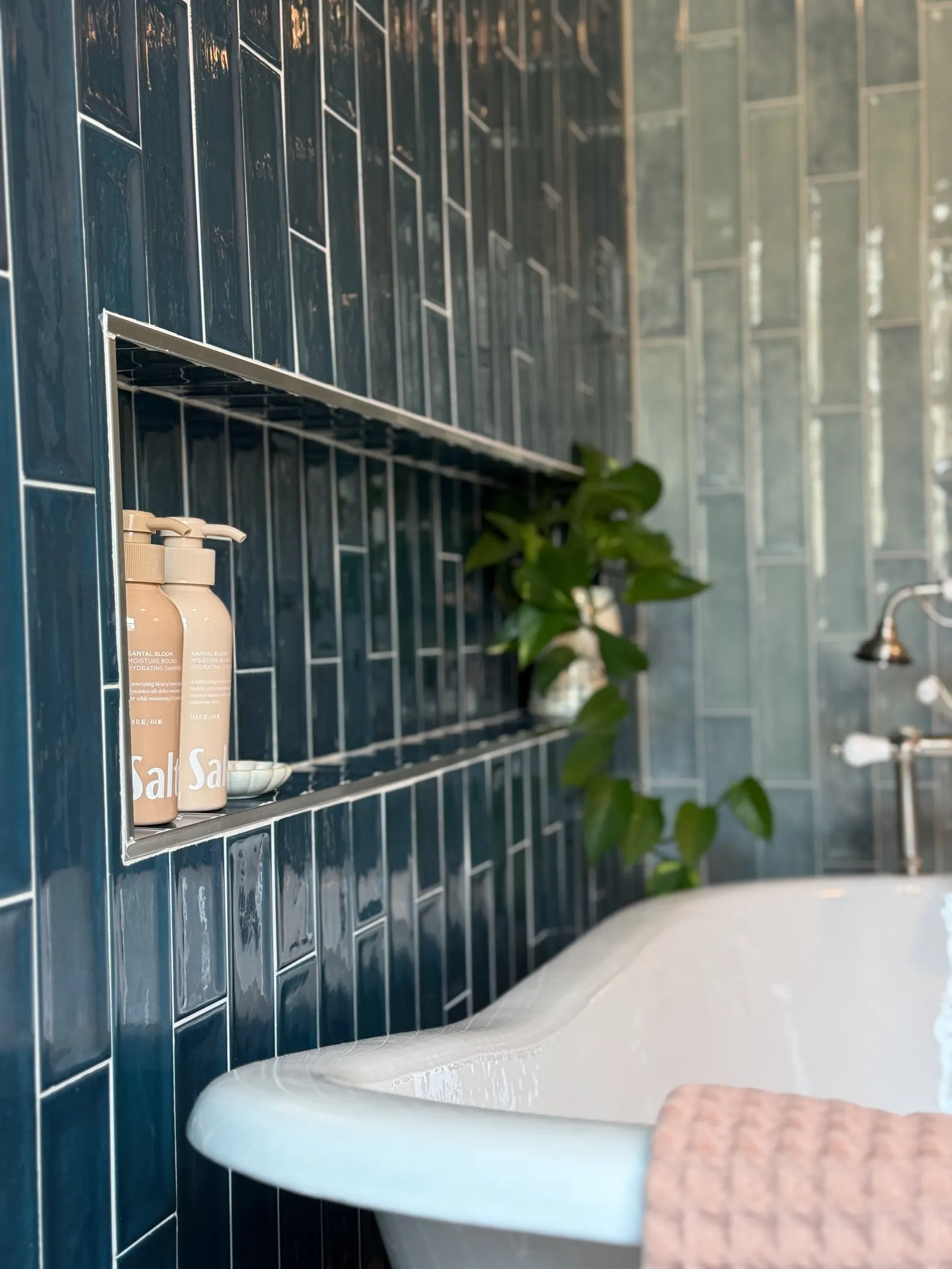 Close-up of a bathroom with dark teal subway tiles, a built-in shelf holding two bottles of moisturizer, a plant, and a white bathtub with a pink towel draped over its edge.