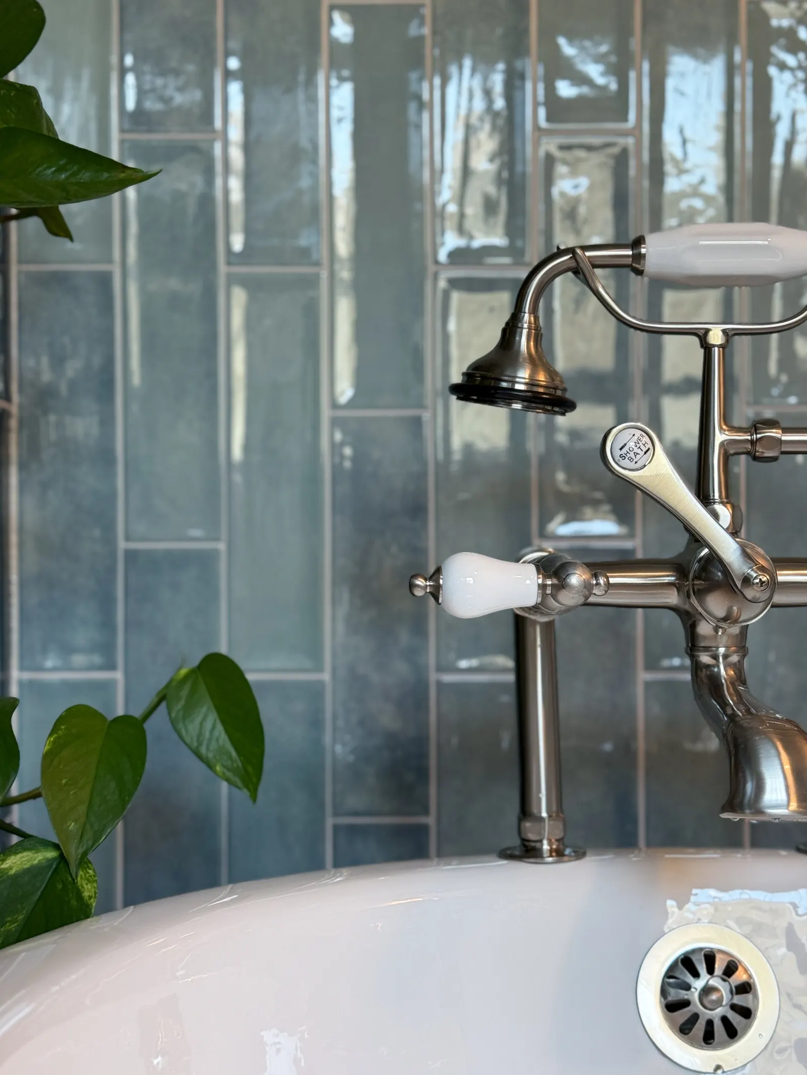 Close-up of a vintage-style bathtub faucet with a showerhead, set against a background of shiny dark gray subway tiles, with green potted plant leaves visible on the left.