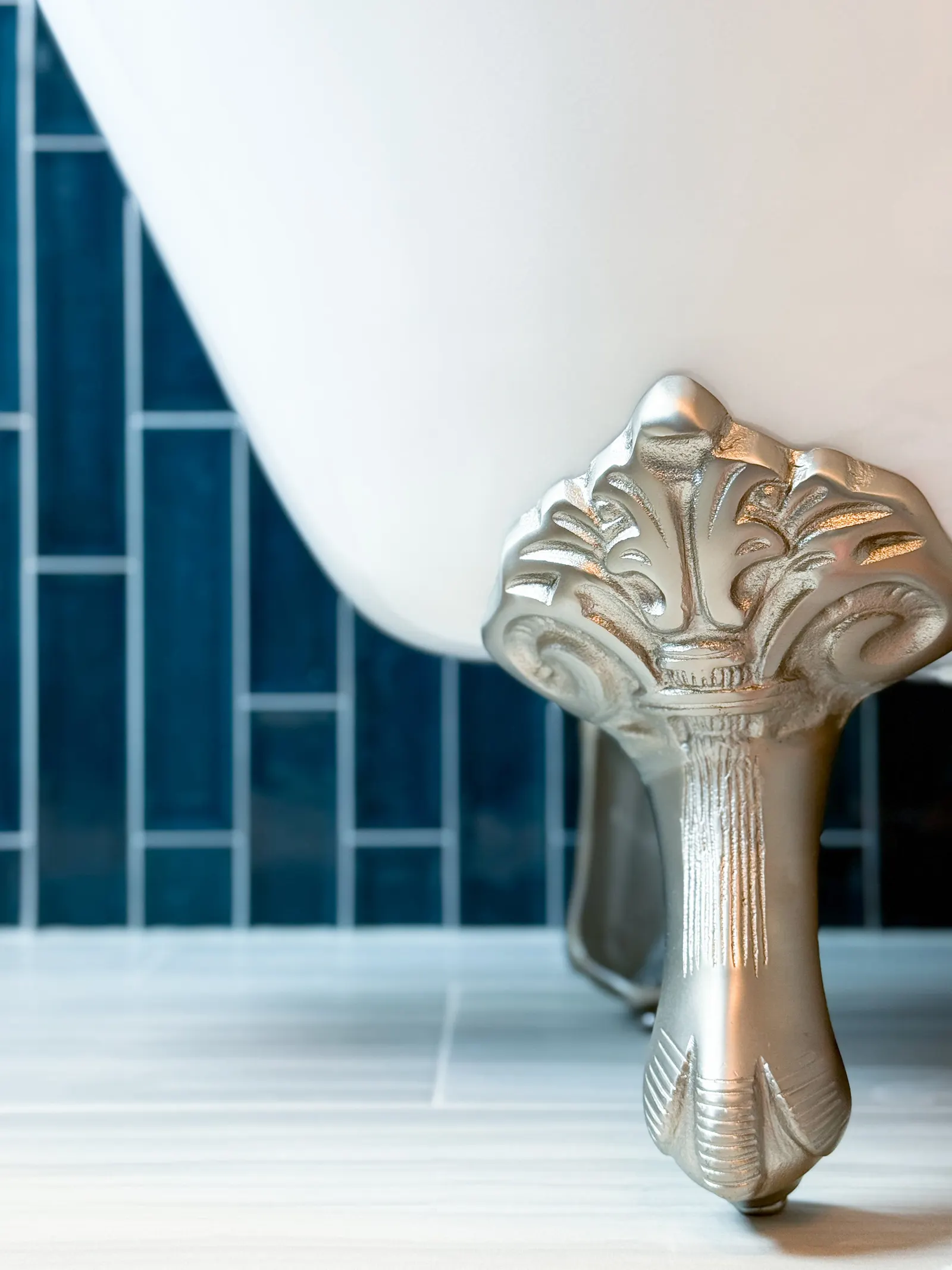 Close-up of a silver clawfoot bathtub leg with ornate detailing, set on a tiled floor with dark blue wall tiles in the background.