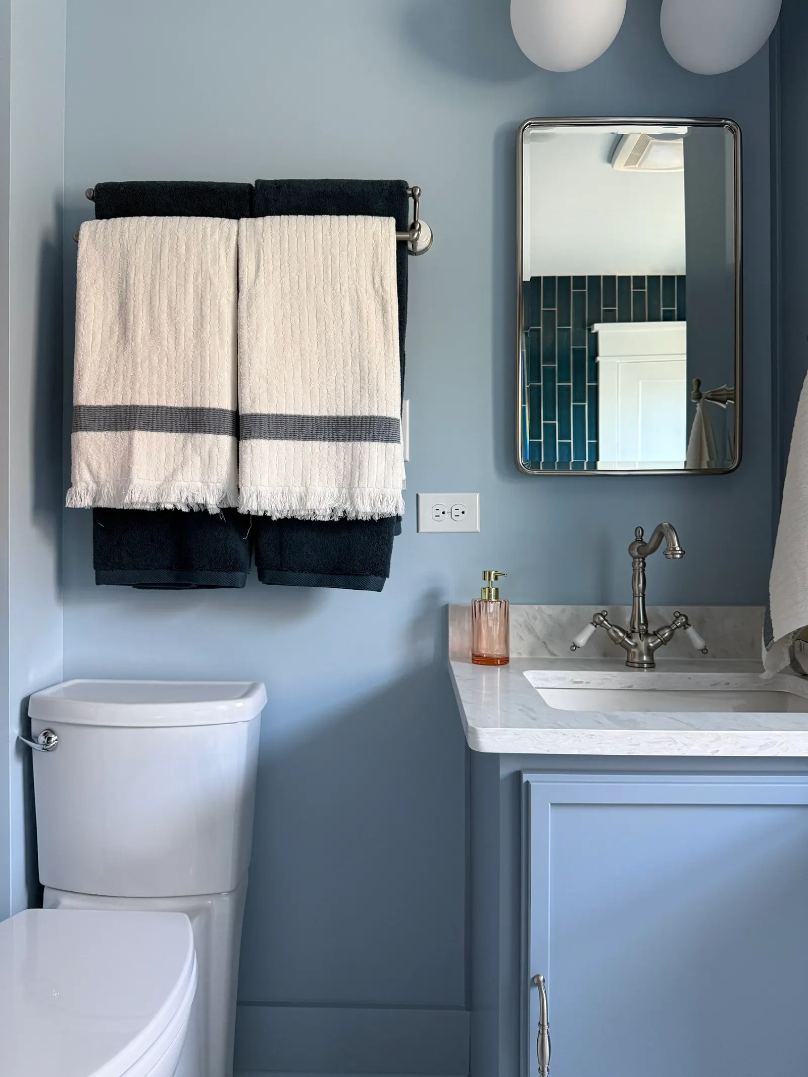 A bathroom with light blue walls, a white toilet, a blue vanity with a white marble countertop, a gold soap dispenser, a rectangular mirror, and a towel rack with black and white towels.