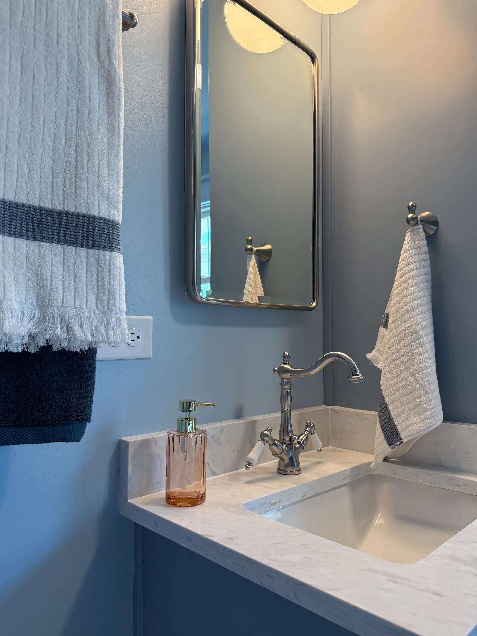 Bathroom sink with marble countertop, a silver faucet, and a pink soap dispenser. Two towels, one white and one dark blue, hanging on silver towel hooks on a blue wall. A rectangular mirror above the sink and a light fixture above the mirror.