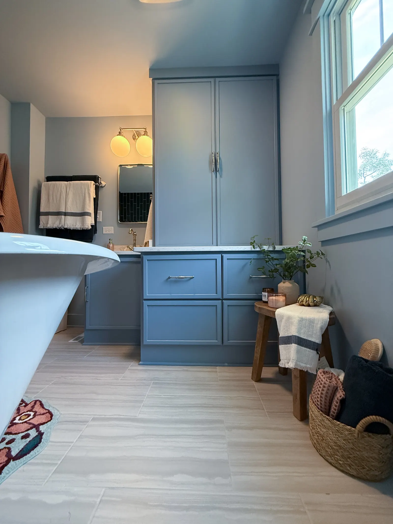 A bathroom with a wooden bench holding a white towel, candles, and a plant near a window. Blue cabinetry and a mirror above the sink are visible, with towels hanging on a rack and a colorful rug on the light-colored tiled floor.