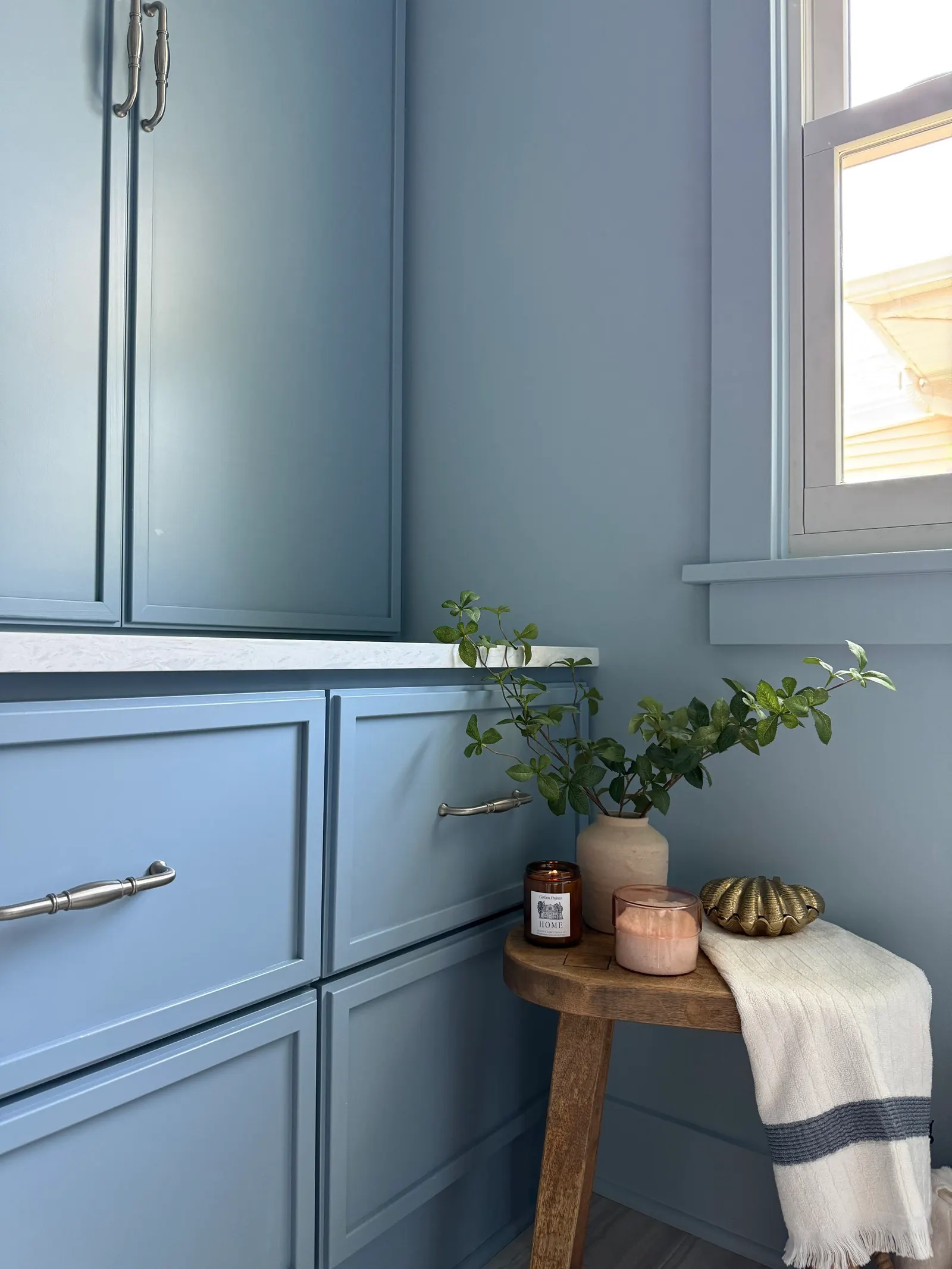 A corner of a room with blue cabinetry, a window, and a small wooden table holding a vase with green foliage, candles, and a decorative gold shell, with a white towel draped over the table.