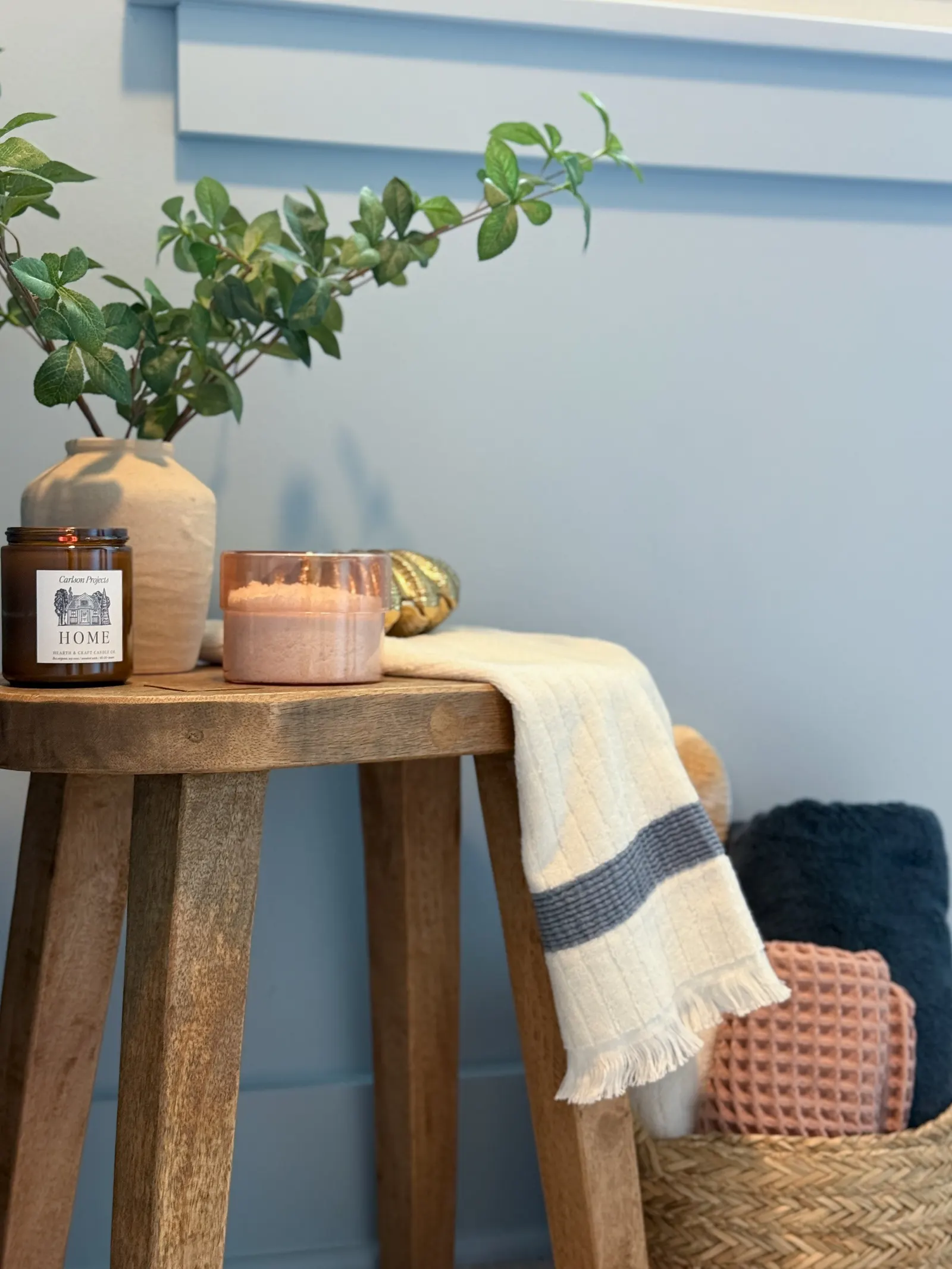 A wooden side table with a beige vase holding green leafy branches, a brown candle, and a pink candle. A white and gray striped cloth drapes over the table's edge. Behind the table, there is a basket with textured pillows—one black, one pink, and one