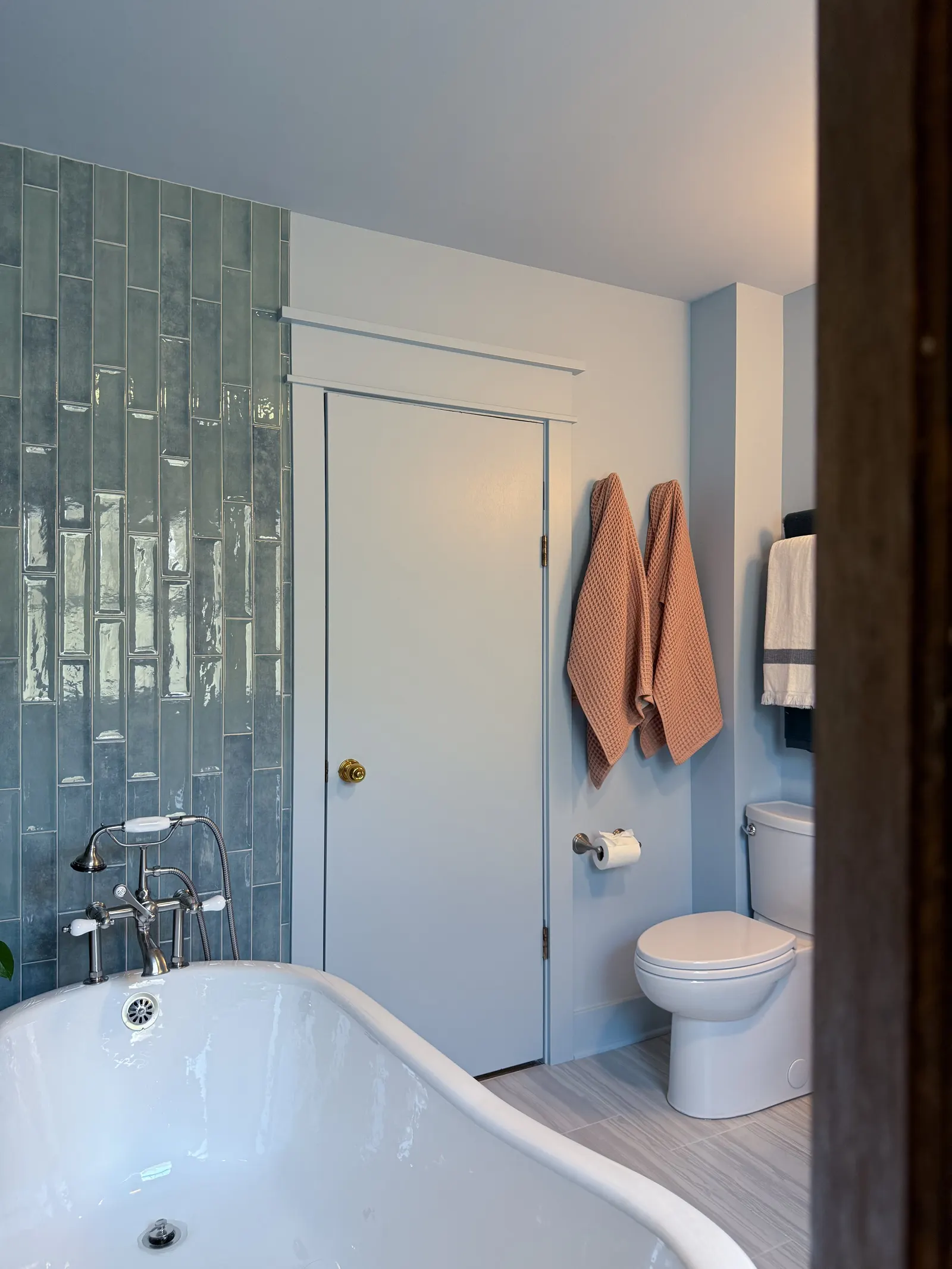 A bathroom with a white bathtub, a toilet, and towels hanging on the wall. The left side features a tiled wall, and there is a door in the center. The bathroom has light-colored flooring and white walls.