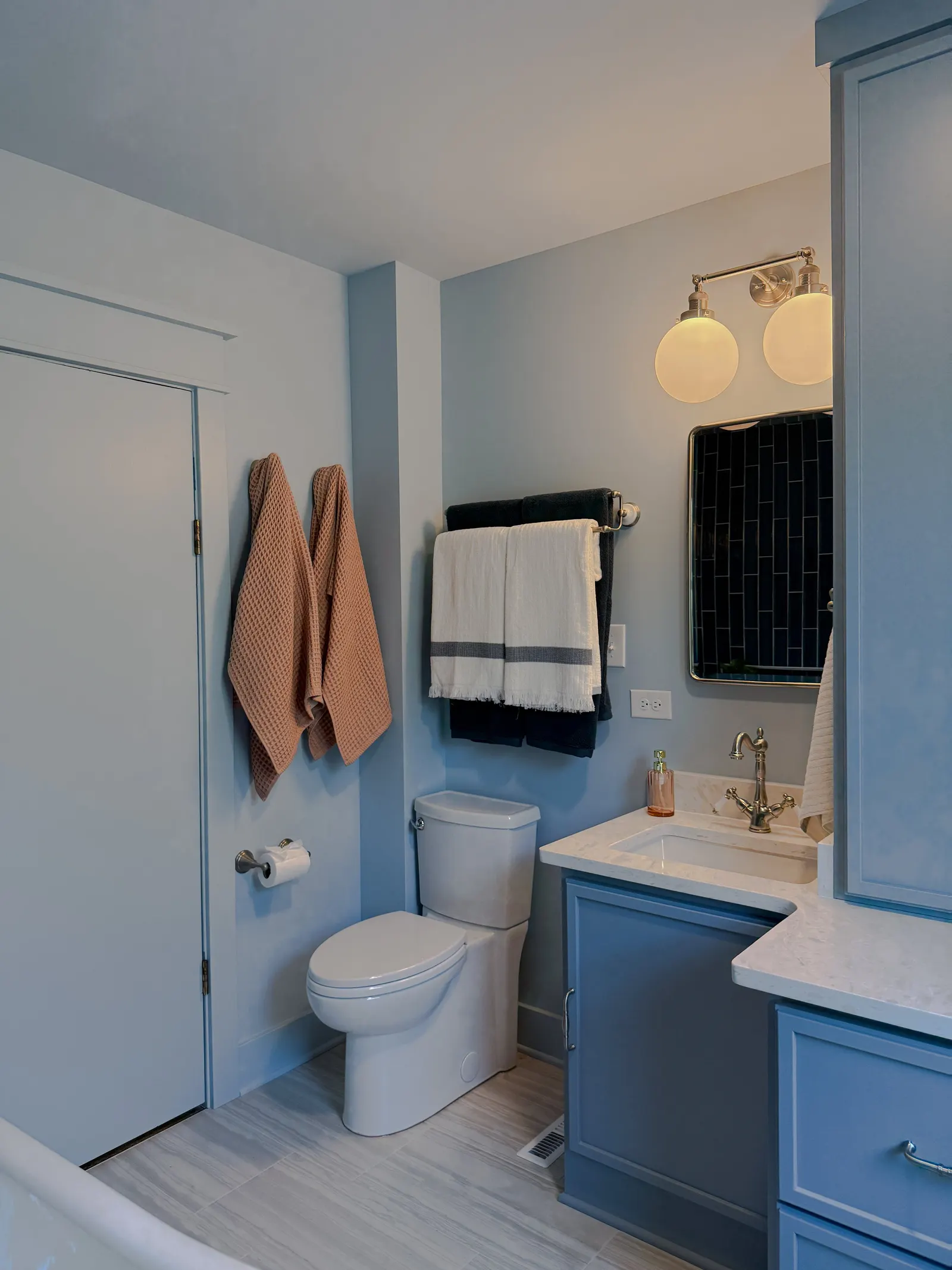 A modern bathroom with light blue walls, a white toilet, a small blue cabinet with a marble countertop, a sink, and a wall-mounted mirror. Two beige towels hang on a towel rack, and two dark towels hang on a second towel rack. Two wall-mounted light