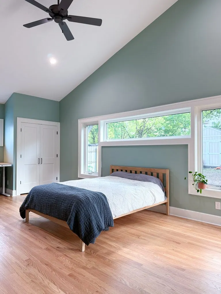 A minimalist bedroom with a wooden bed frame, white bedding, a dark blue quilt, and a gray pillow. The room has light green walls, hardwood flooring, and three large windows with a view of trees and a fence outside. There is a small potted plant on t