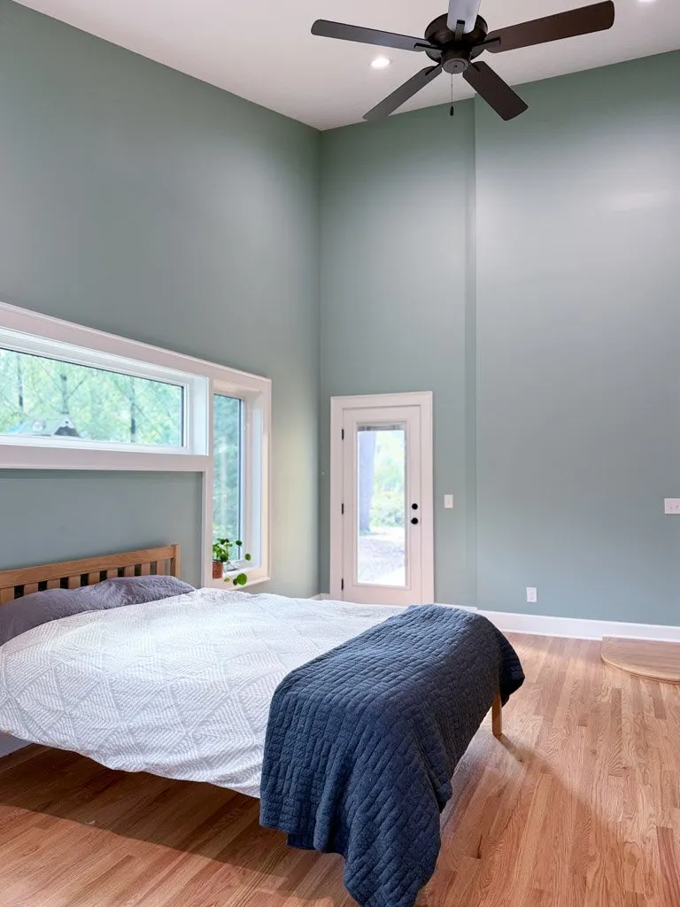 A bedroom with light green walls, a wooden bed frame with white bedding, a dark blue quilt, a window, a glass door, a ceiling fan, and hardwood floors.