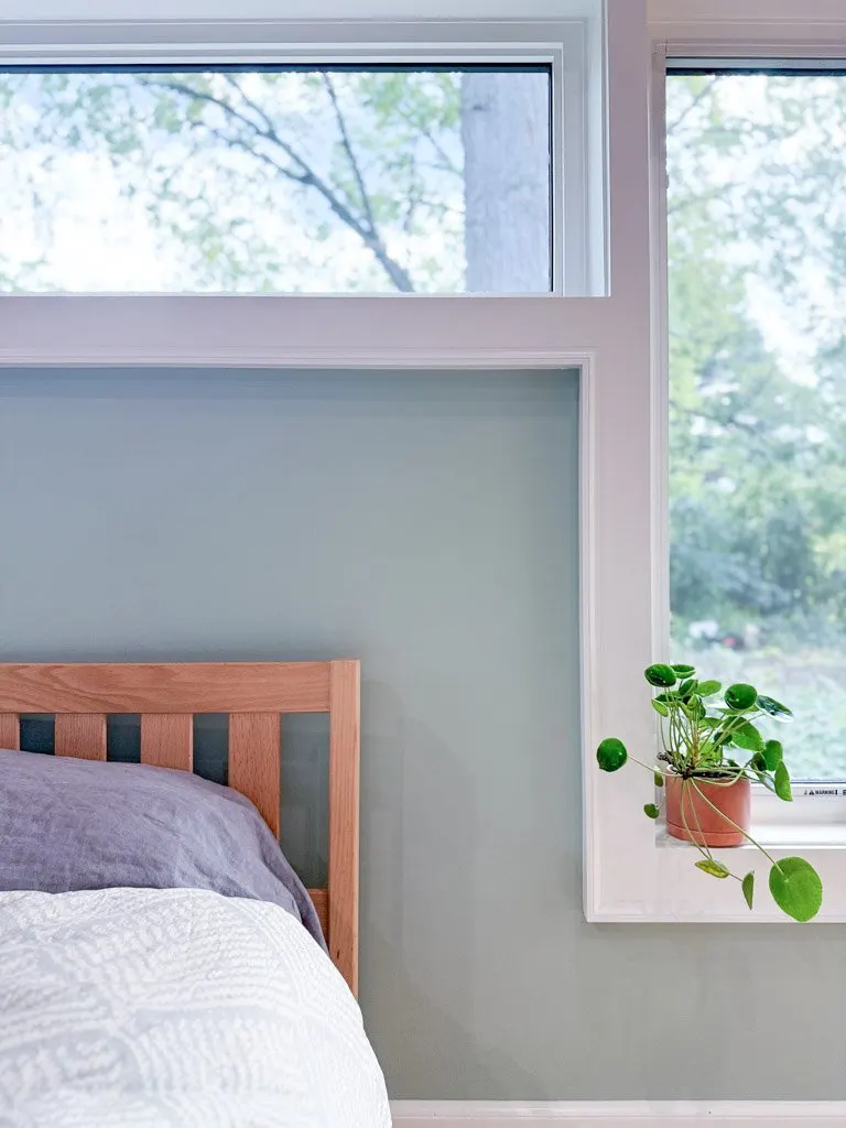 Interior view of a bedroom corner with a wooden bed frame, white and purple bedding, and a window with a plant on the windowsill.