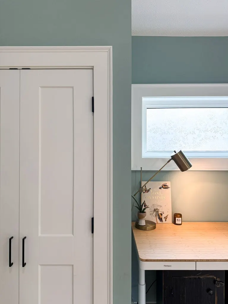 A corner of a room with double white closet doors on the left and a small wooden desk with a brass desk lamp, a potted plant, a decorative book, and a candle on it, situated under a window with frosted glass.