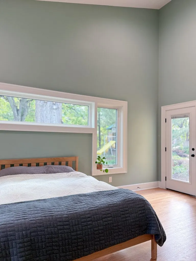 A bedroom with light green walls, a bed with a wooden frame, white and gray bedding, and a large window showing outdoor greenery.