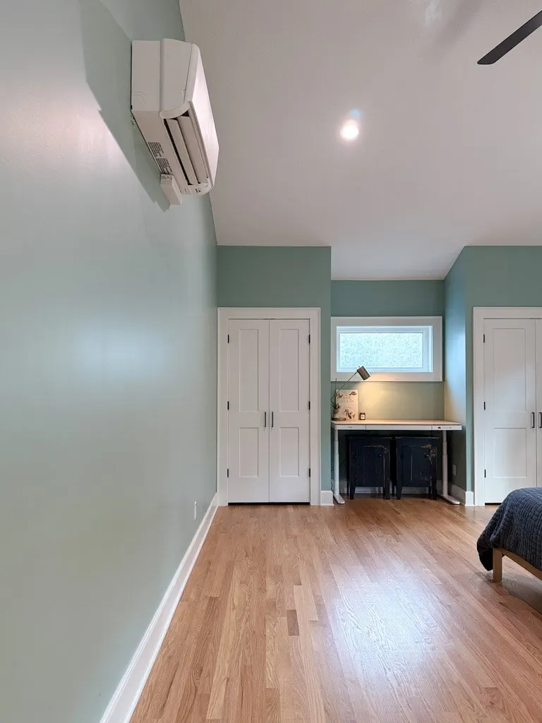 A room with light green walls, hardwood floor, and a white ceiling. There is an air conditioning unit mounted on the wall, a window with frosted glass, a small desk with a lamp, and a bed partially visible on the right side.