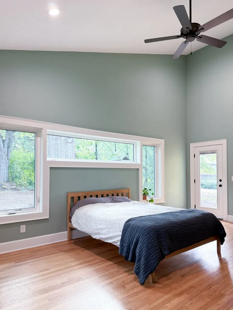 Empty bedroom with light green walls, a wooden bed frame with a white quilt and dark blue blanket, large windows, a potted plant, and a ceiling fan.