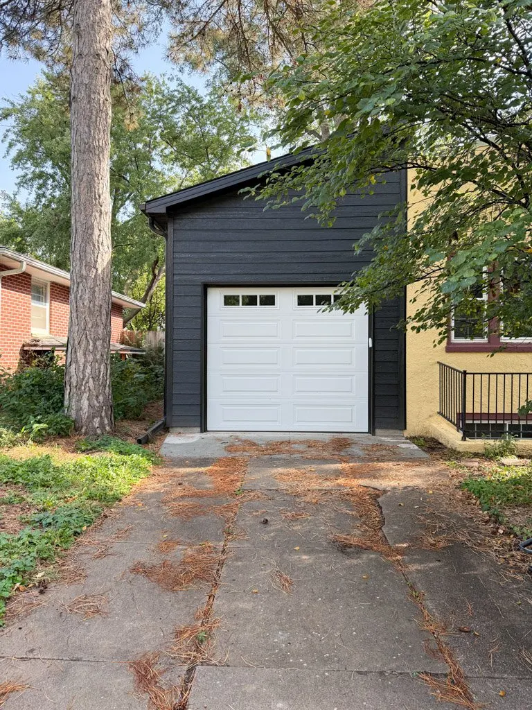 A residential garage with a white door, black wooden siding, and a concrete driveway covered with pine needles. There is a large tree on the left and a small tree with green leaves on the right, with neighboring houses partially visible.