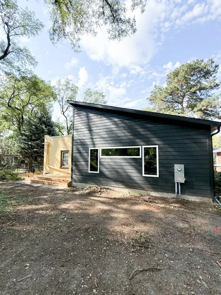 A modern house with black wooden siding and a unique sloped roof, featuring two rectangular windows, an electrical meter on the side, and a small deck with steps, set among trees with a partly cloudy sky overhead.