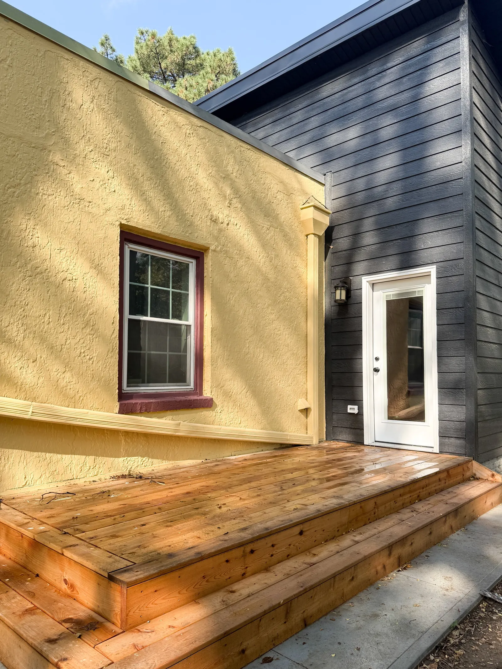 Backyard deck under construction with wooden platform, yellow stucco and black siding house exterior, window, and glass door.
