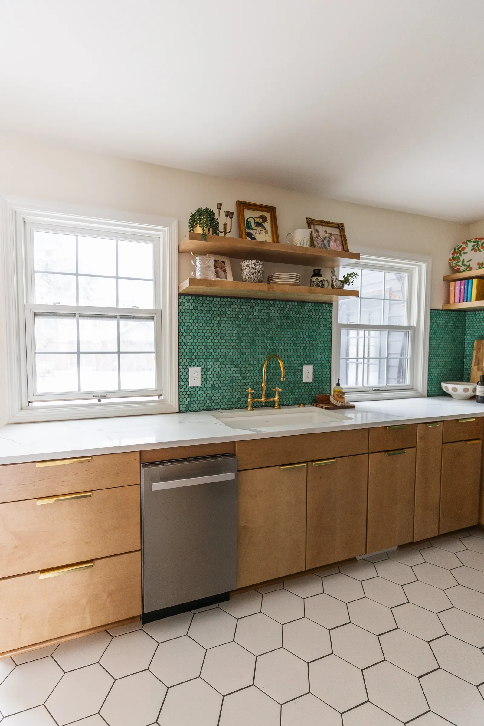 Modern kitchen with two windows, wooden cabinets with gold handles, a white countertop, green hexagonal tile backsplash, gold faucet, open shelves with framed photos, plants, and decorative items, and white hexagonal floor tiles.