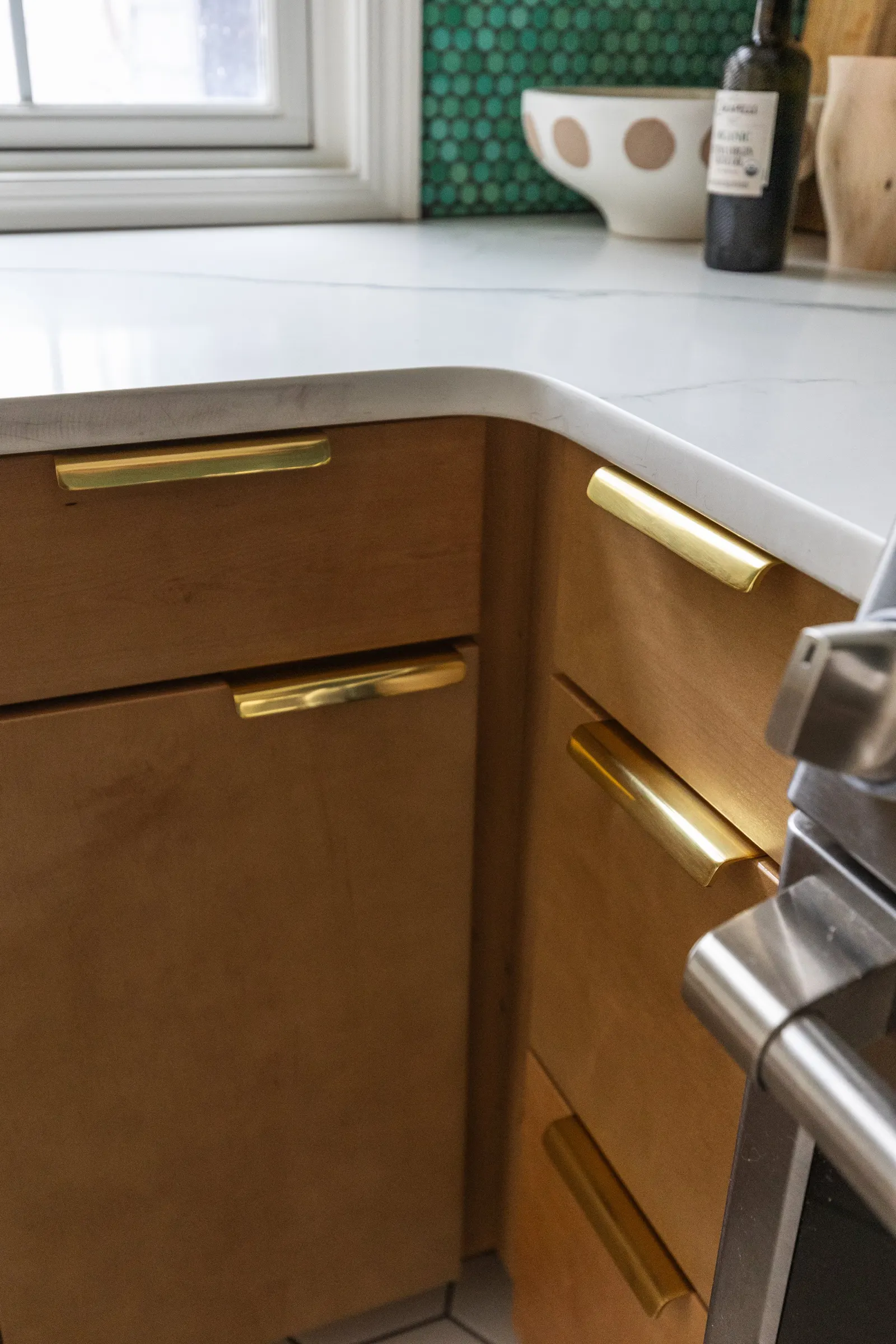 Close-up of a kitchen corner with wooden cabinets with gold handles and a white marble countertop, with items on the counter including a bowl, a bottle, and a window in the background.