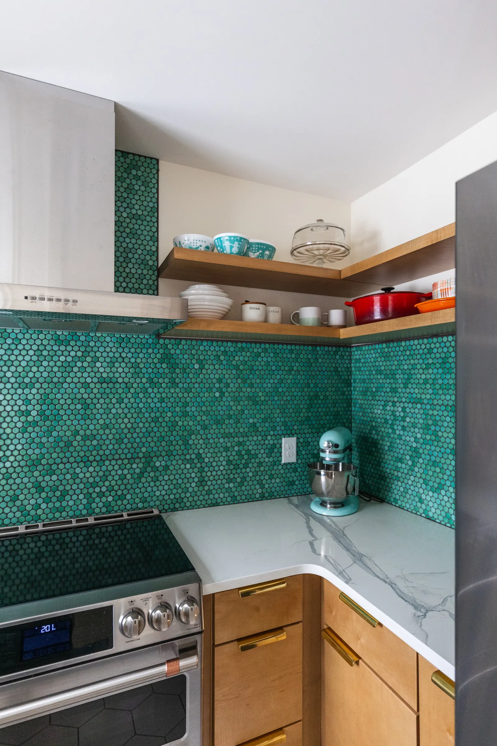 Corner of a modern kitchen with green hexagonal tile backsplash, wooden shelves holding dishes and cookware, and a white marble countertop with teal mixer