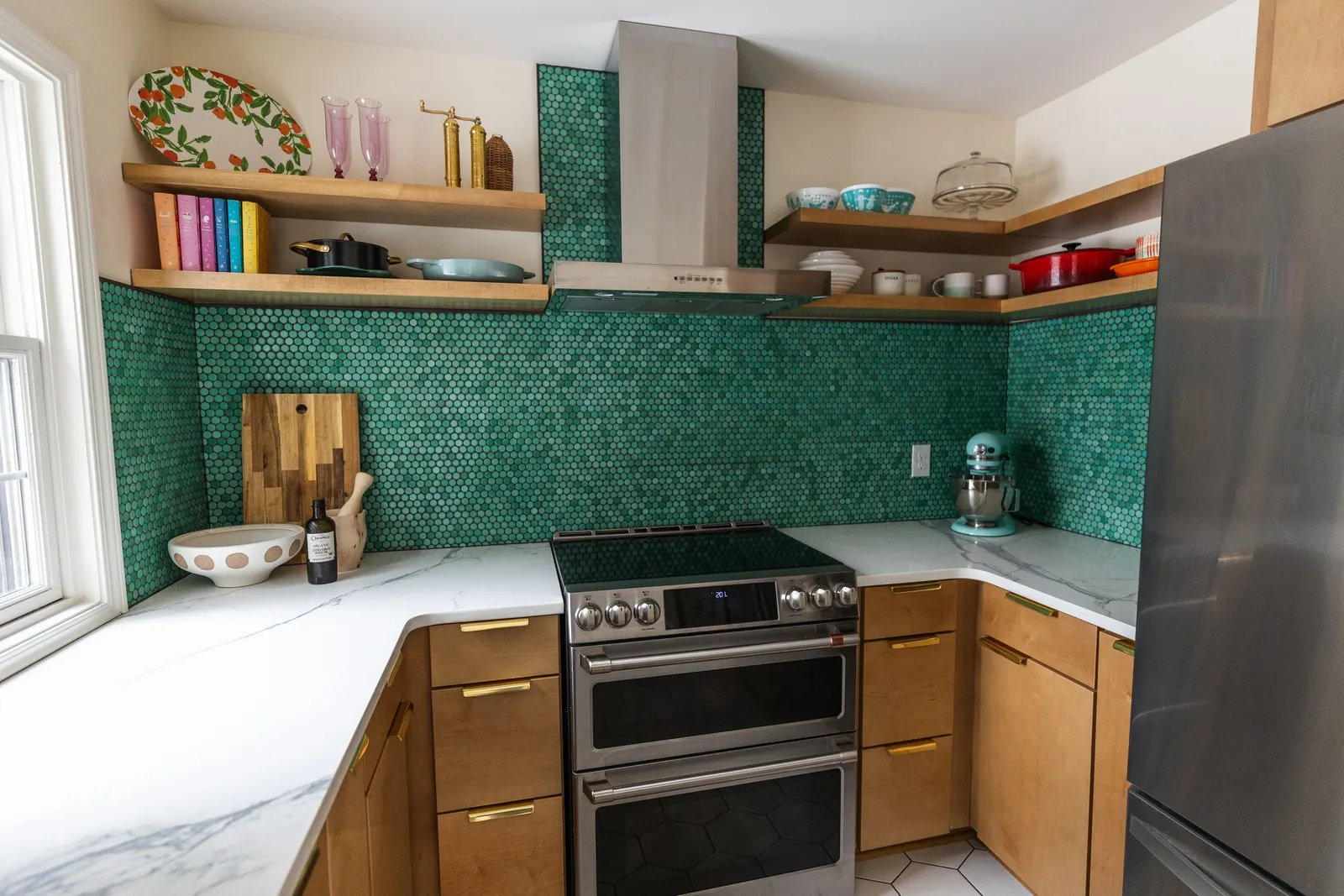 Kitchen with green hexagonal tile backsplash, wooden cabinets with gold handles, marble countertop, stainless steel double oven, open shelving with colorful dishes, cookbooks, and kitchen decor.
