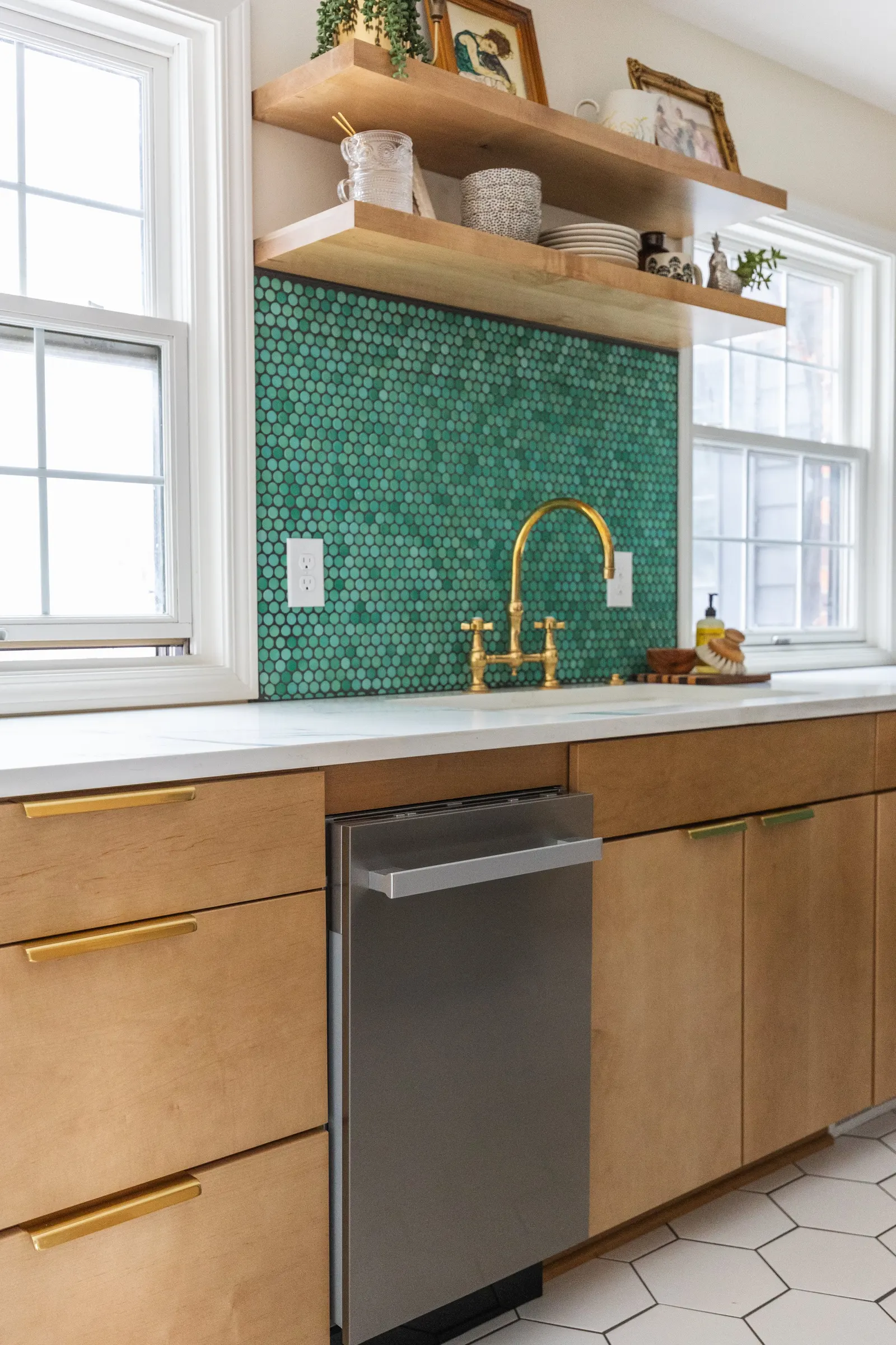 Kitchen with wooden cabinets, a white marble countertop, green hexagonal tile backsplash, and a gold faucet. Open wooden shelves above display decorative items, framed artwork, and potted plants. Two windows let in natural light.