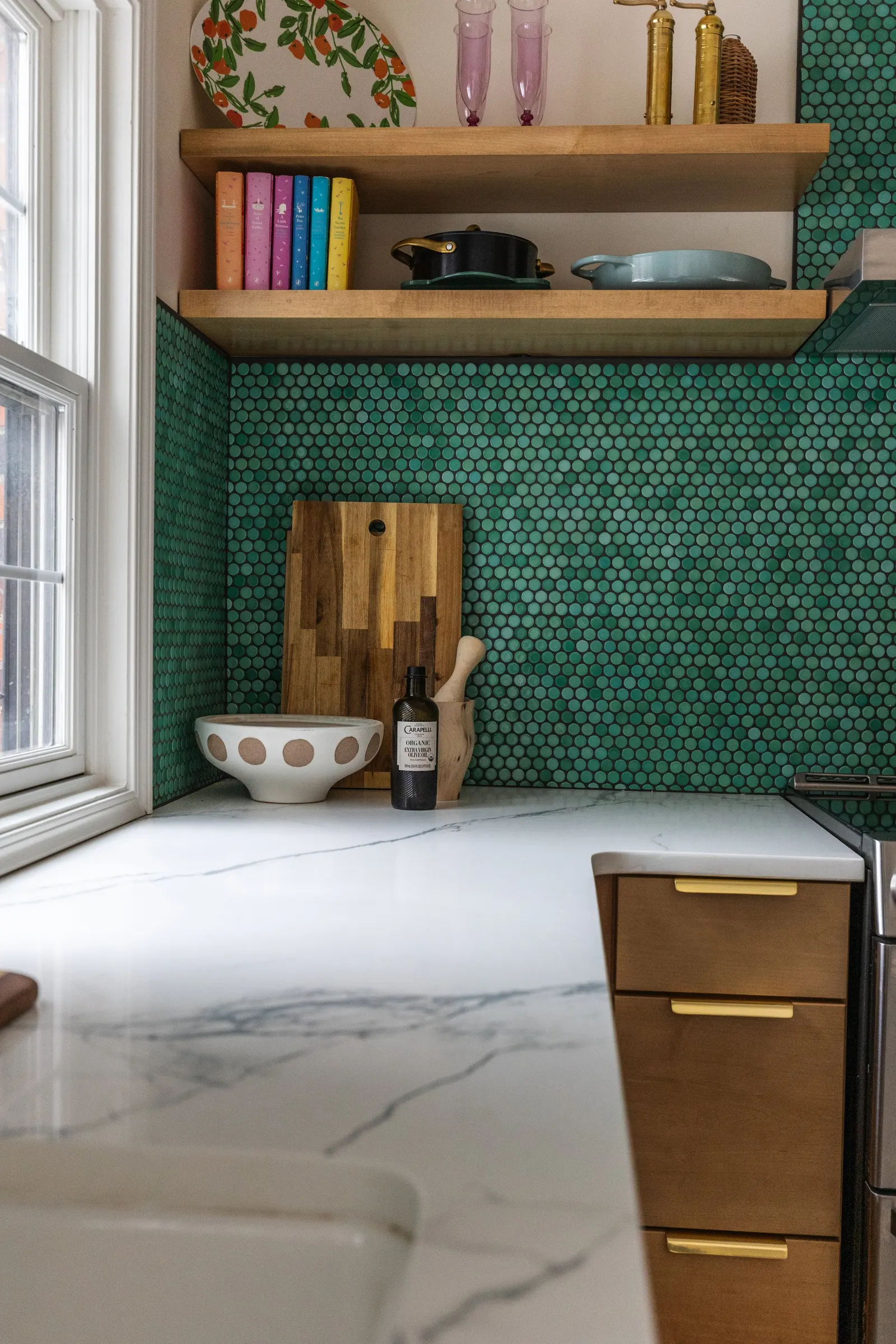 A modern kitchen with green hexagonal tile backsplash, wooden open shelves, a marble countertop, and decorative items including a wooden cutting board, a bowl, a soap dispenser, and some books and glassware.