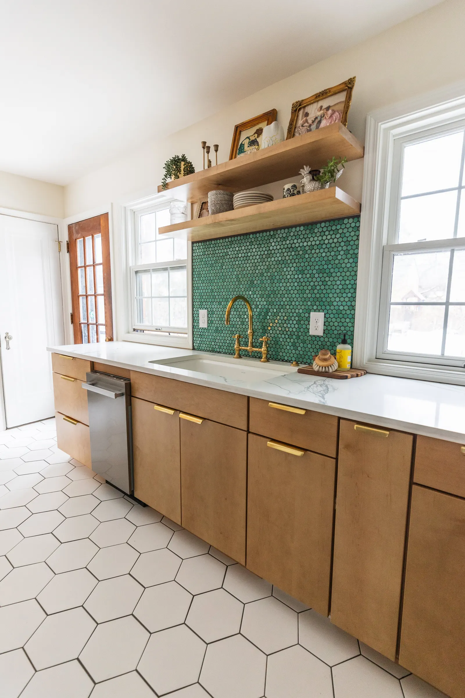 Kitchen with wooden cabinets, white marble countertop, green hexagonal tile backsplash, gold faucet, and open wooden shelves with decorative items. Two windows and a wooden door are visible.