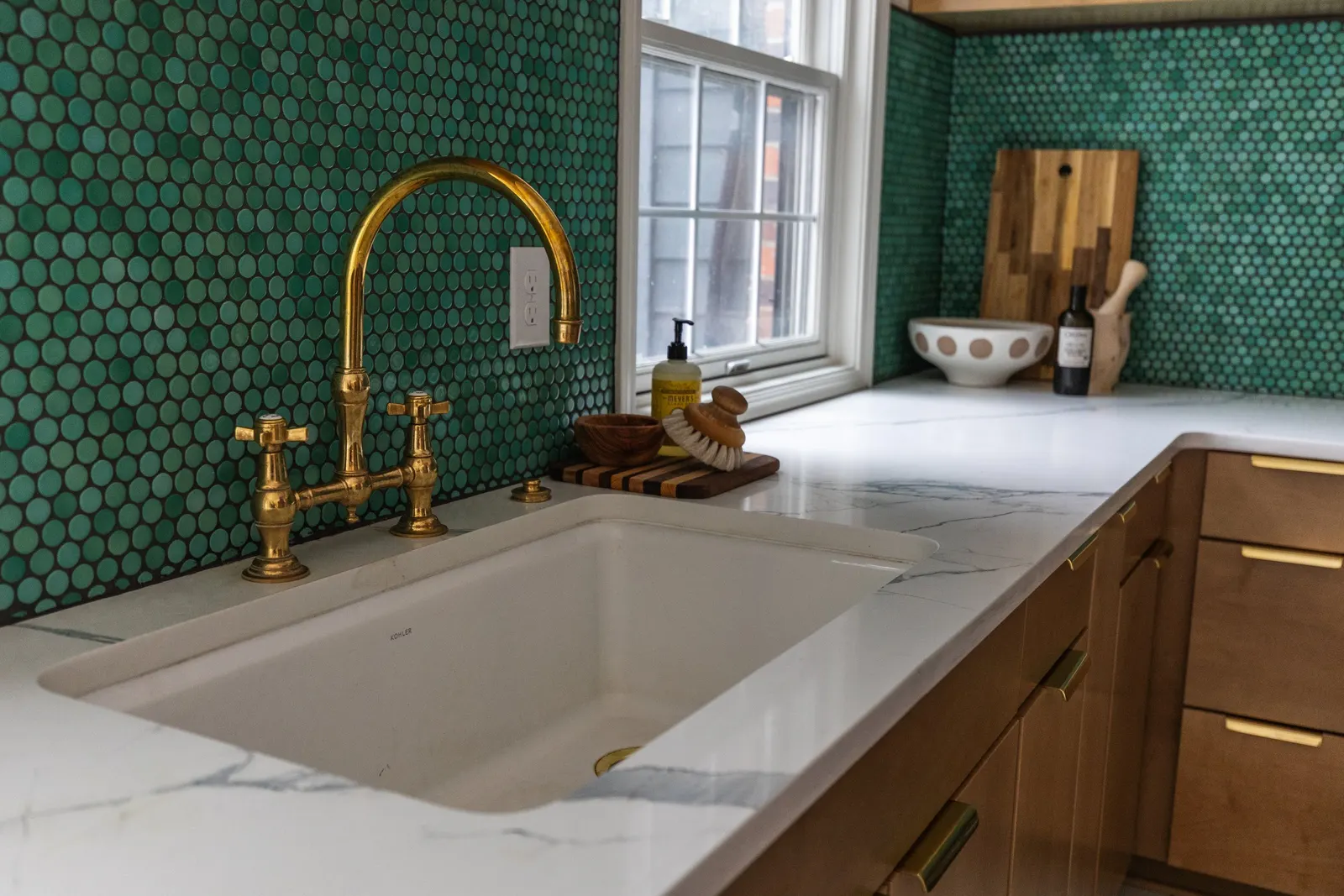 A kitchen sink with a gold faucet and handles, a white marble countertop, a green dotted tile backsplash, and a window with a view outside. Items on the counter include a soap pump, wooden bowl, cleaning brush, and decorative objects.